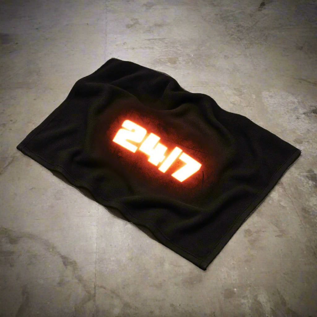 Black rally towel displayed on a concrete floor with visible folds, featuring a bold white '24/7' graphic at the center, glowing with a fiery red-orange effect around the text.
