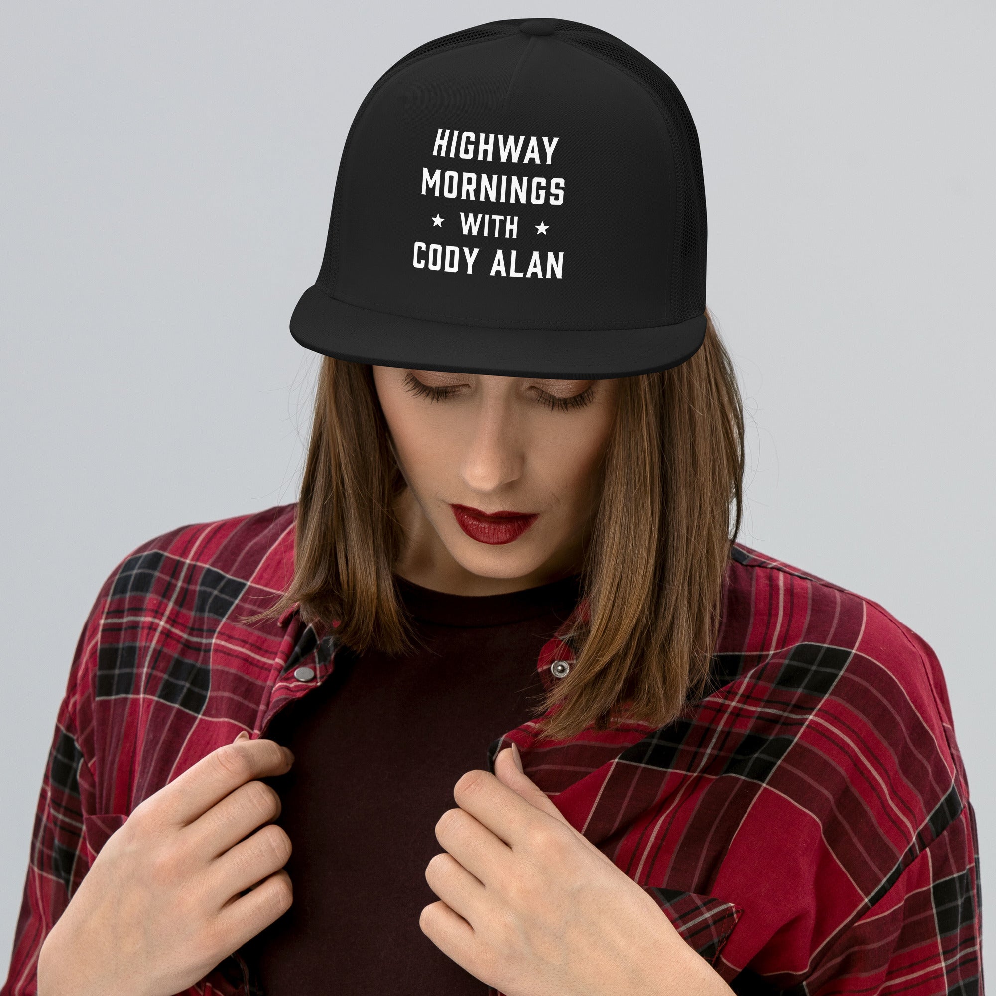 A woman wearing a black trucker hat with the phrase 'HIGHWAY MORNINGS WITH CODY ALAN' on the front. She is looking down and adjusting her red plaid flannel shirt over a dark top.