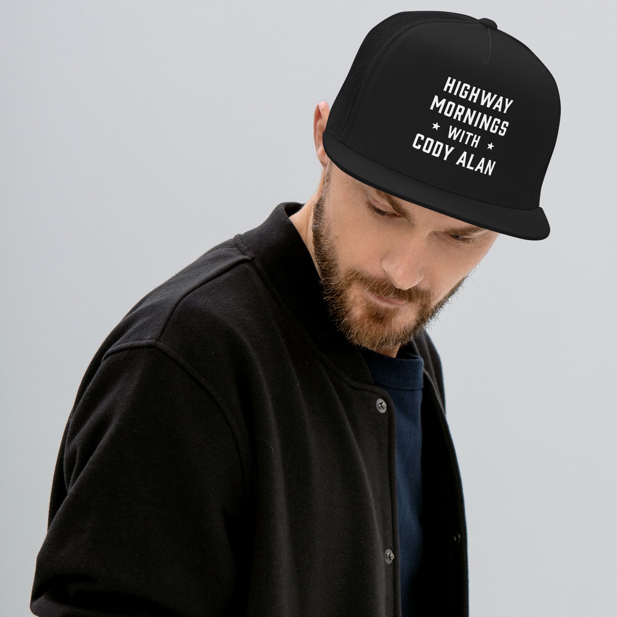 A man looking down while wearing a black trucker hat with white text on the front that reads 'HIGHWAY MORNINGS WITH CODY ALAN'. He is dressed in a black jacket and dark shirt.