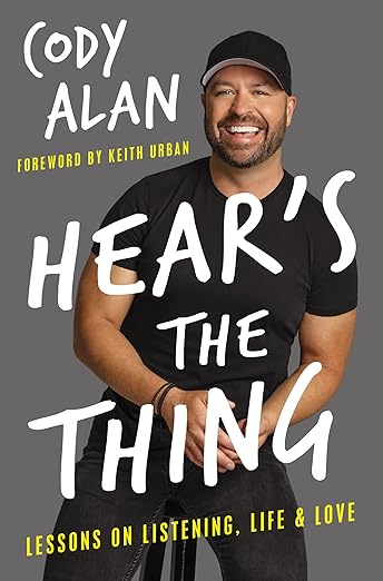 The front book cover of 'Hear’s the Thing by Cody Alan,' featuring a smiling Cody in a black shirt and hat against a gray background with large white handwritten-style text and yellow subtext reading 'Lessons on Listening, Life & Love' and a foreword credit to Keith Urban.