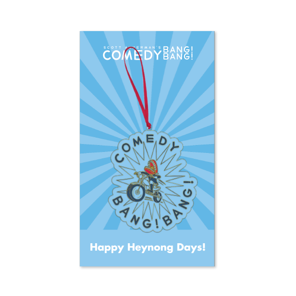 Blue backer card with rays radiating from the center and white text reading ‘Scott Aukerman’s Comedy Bang! Bang!’ at the top. The ornament attached shows a person on a motorcycle with the words ‘COMEDY BANG! BANG!’ around the design, hanging from a red ribbon. Text at the bottom reads ‘Happy Heynong Days!’