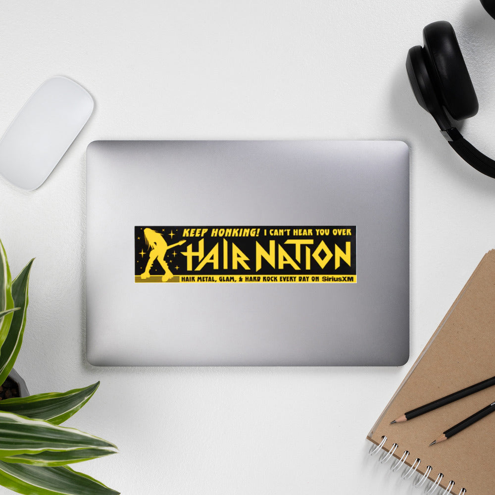 A laptop with a black sticker with yellow text that says 'KEEP HONKING! I CAN'T HEAR YOU OVER HAIR NATION' featuring hair metal themes.