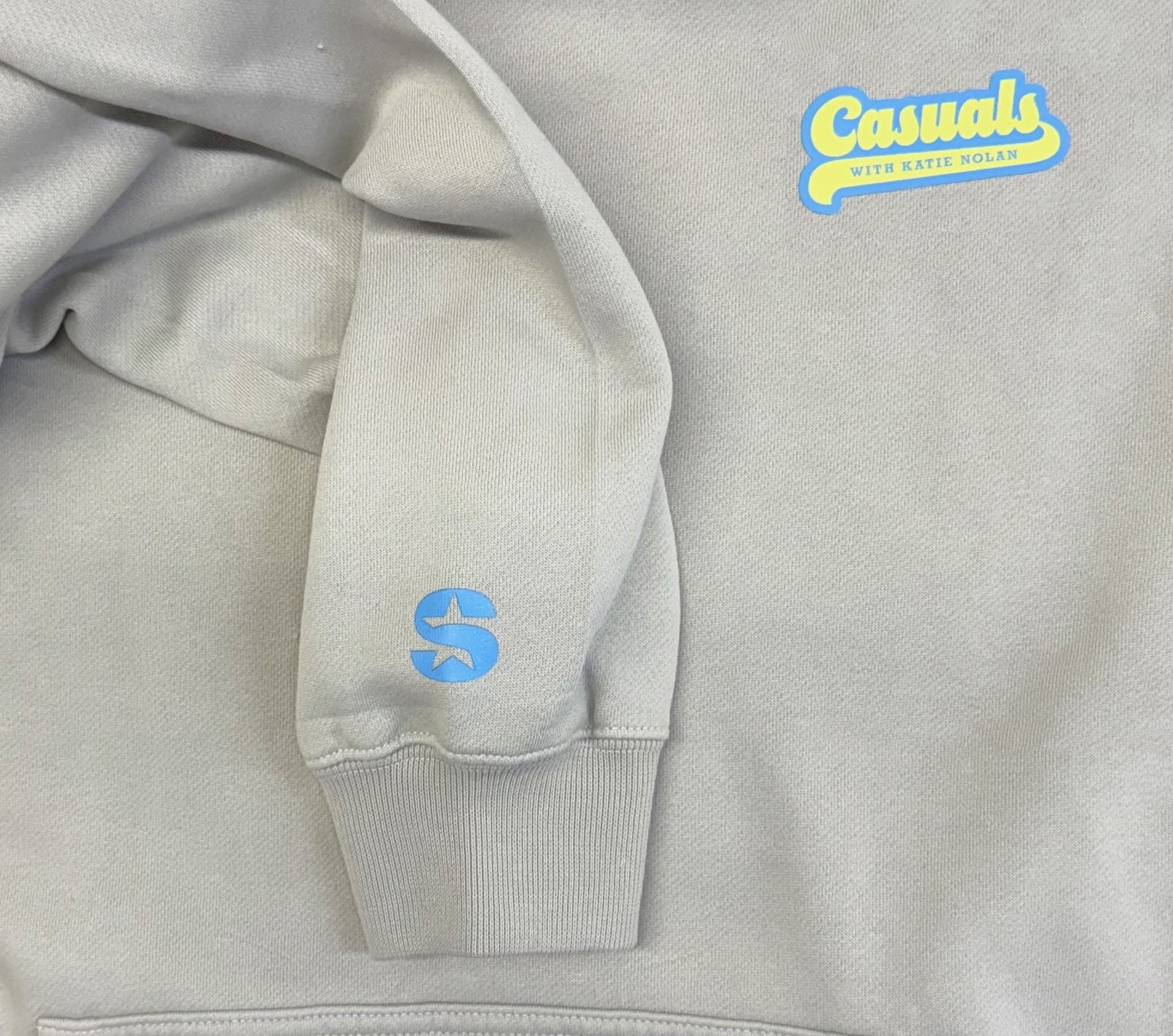 Detail view of an off-white hoodie with blue and yellow ‘Casuals with Katie Nolan’ logo printed on the left chest and a small blue SiriusXM S-Star logo on the left wrist cuff.