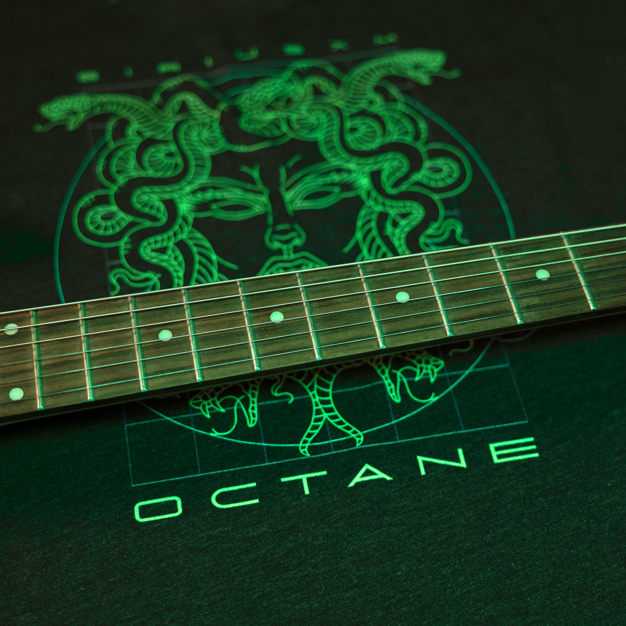 Guitar neck resting on a dark t-shirt with a glowing green Medusa design and the word 'OCTANE.'