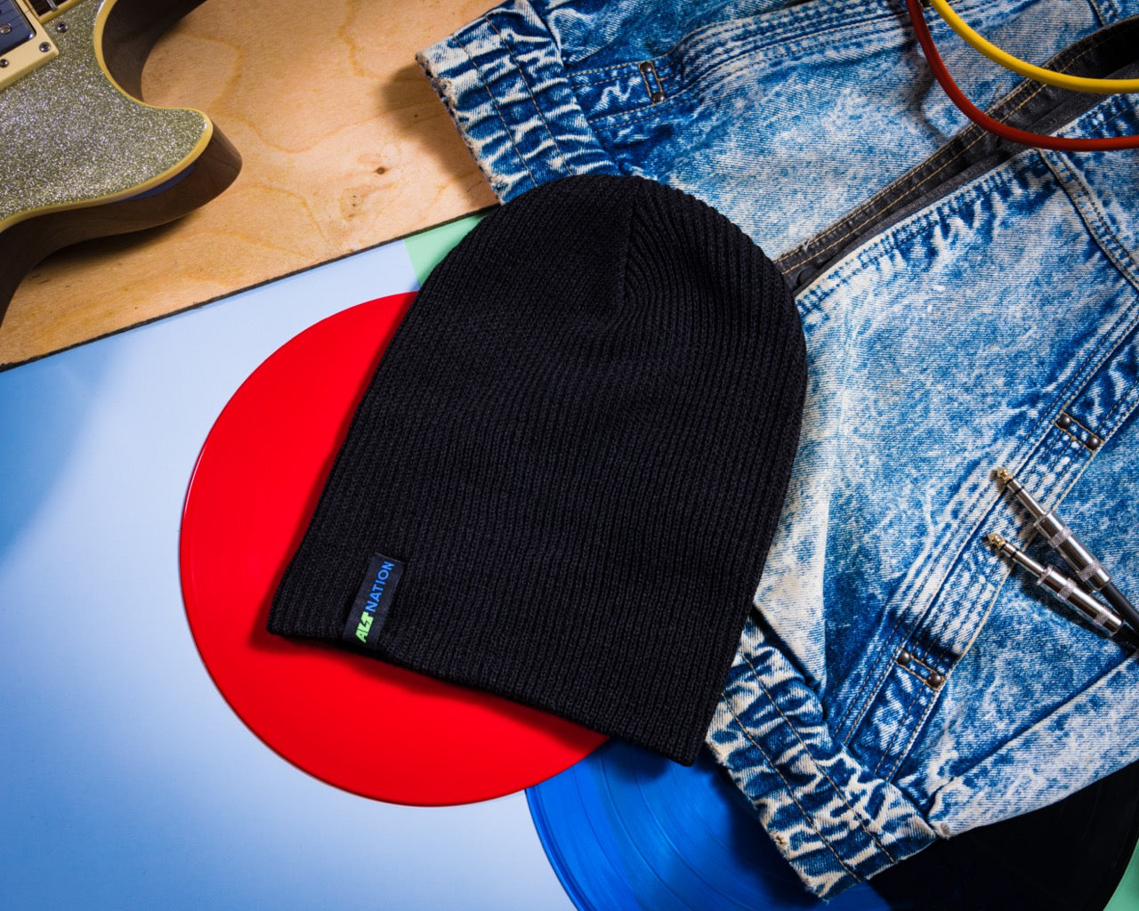 Black ribbed 'alt nation' tagged beanie placed on a red vinyl record, surrounded by denim fabric and a sparkly guitar.
