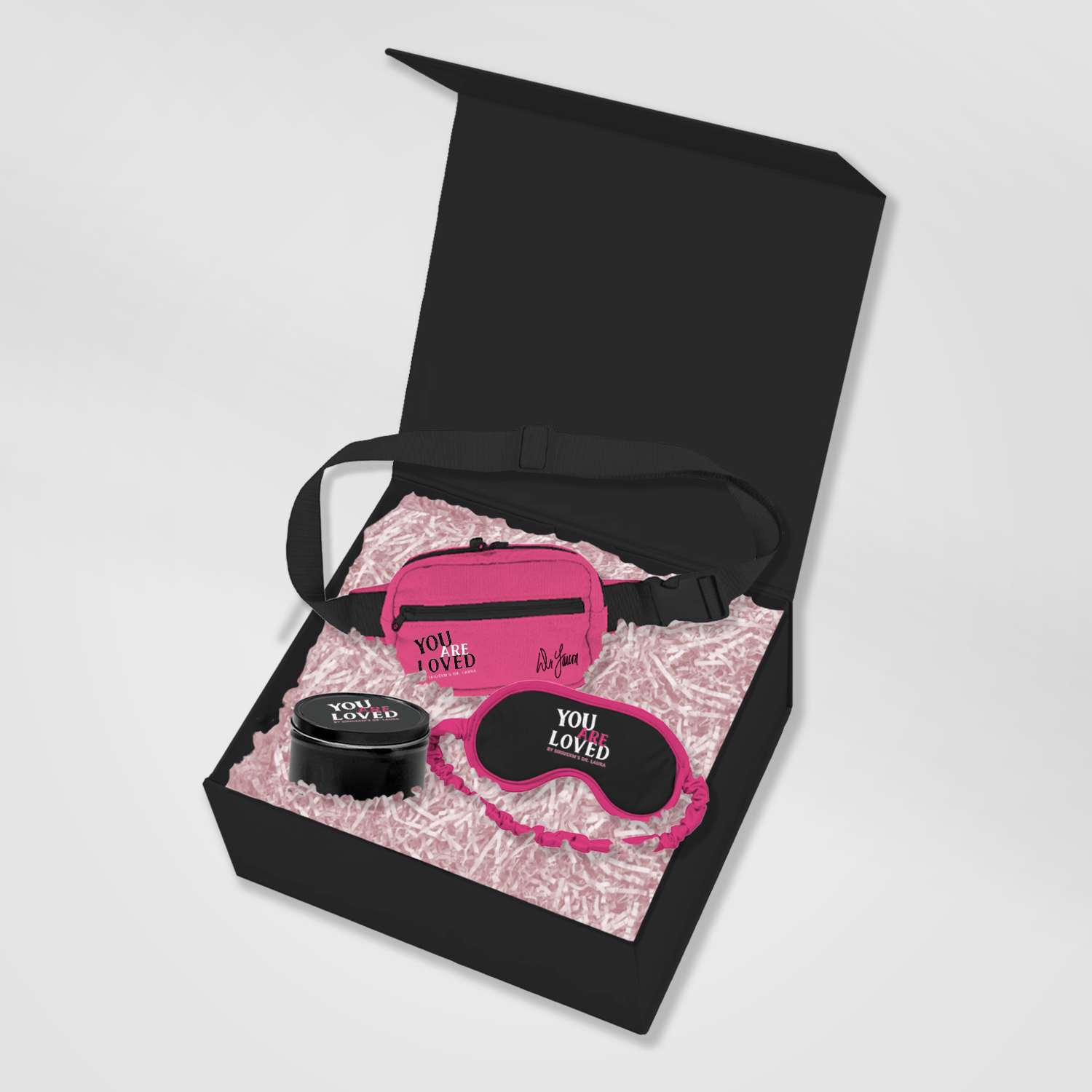 An open black gift box filled with pink crinkle paper, containing a pink hip pack, a black and pink sleep mask, and a black candle tin—each printed with the phrase ‘You Are Loved.’