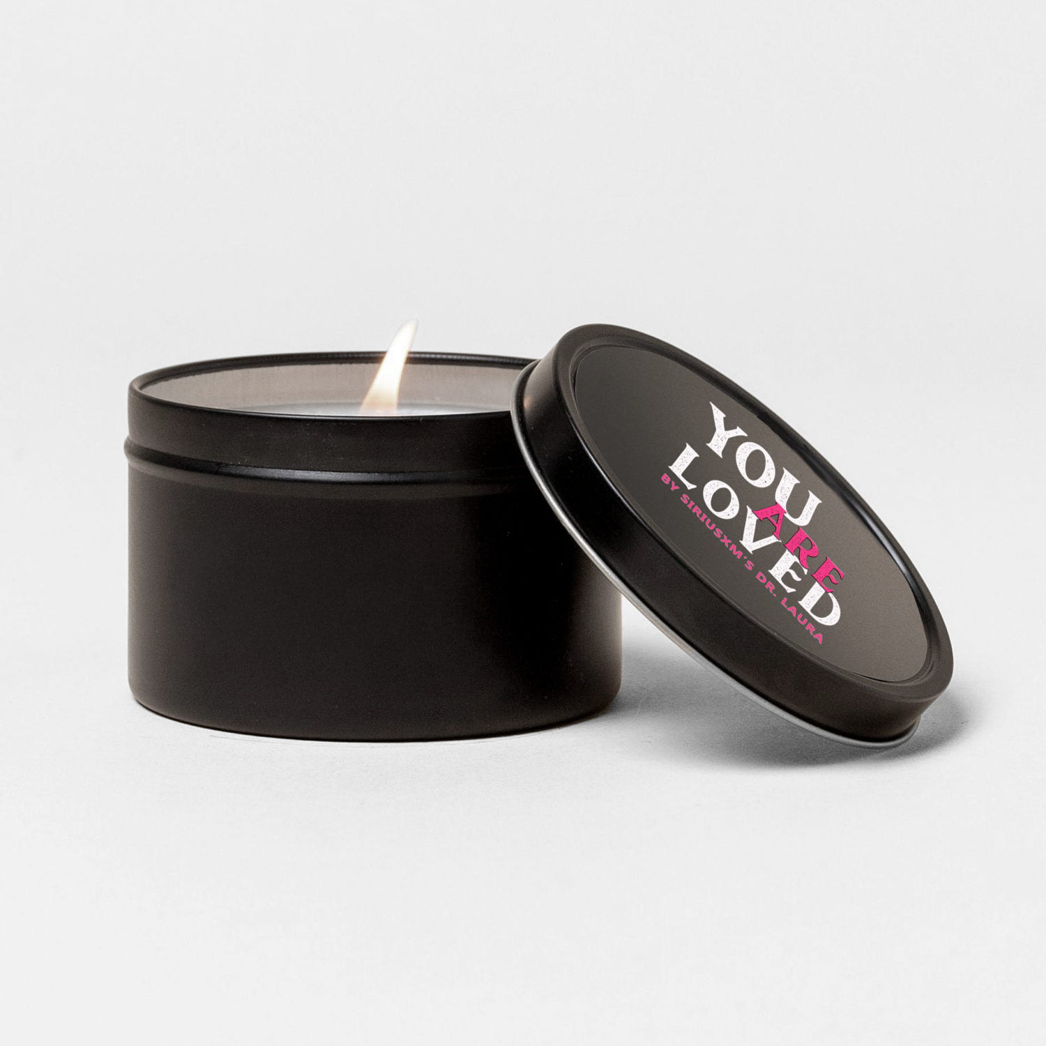 A small black candle tin with its lid slightly open to reveal a lit candle. The lid is labeled ‘You Are Loved by SiriusXM’s Dr. Laura’ in white and pink text.