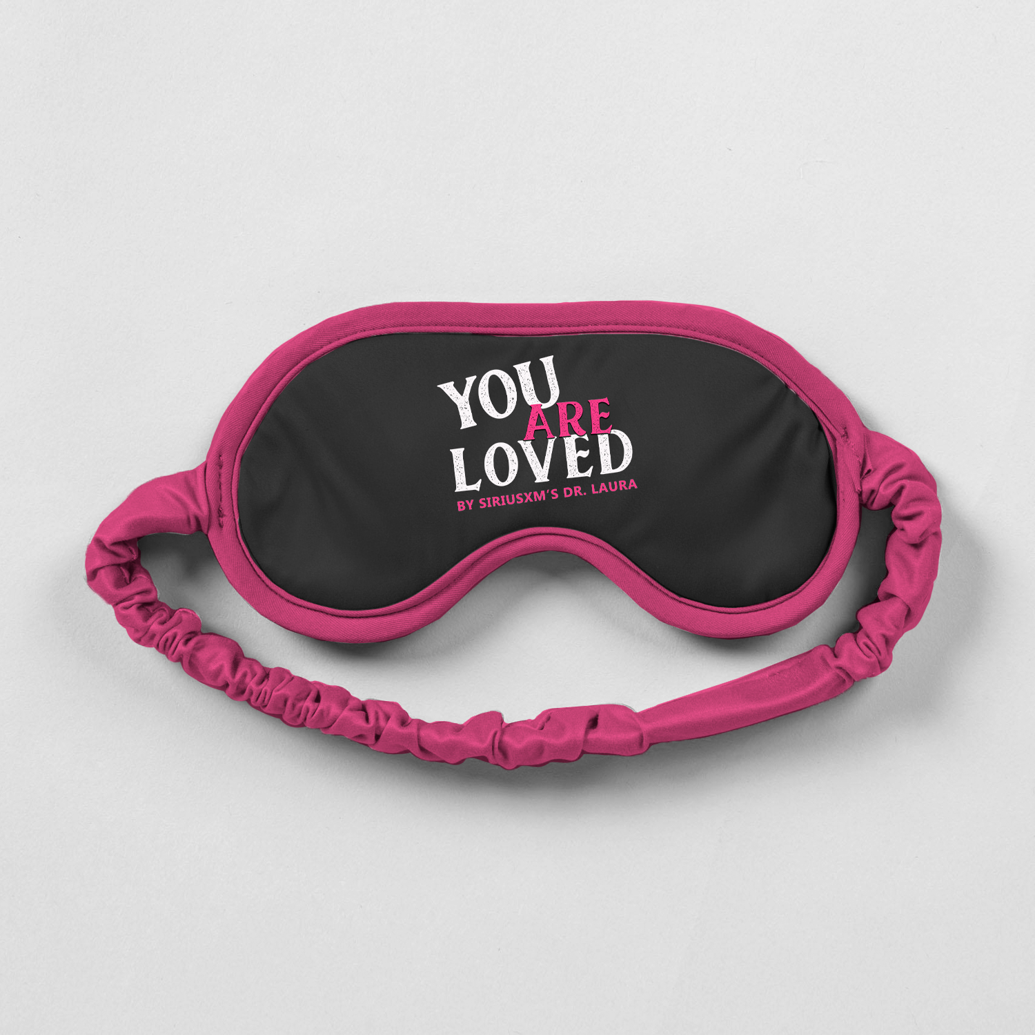 A black sleep mask with pink trim and strap, displaying the phrase ‘You Are Loved by SiriusXM’s Dr. Laura’ in white and pink text.