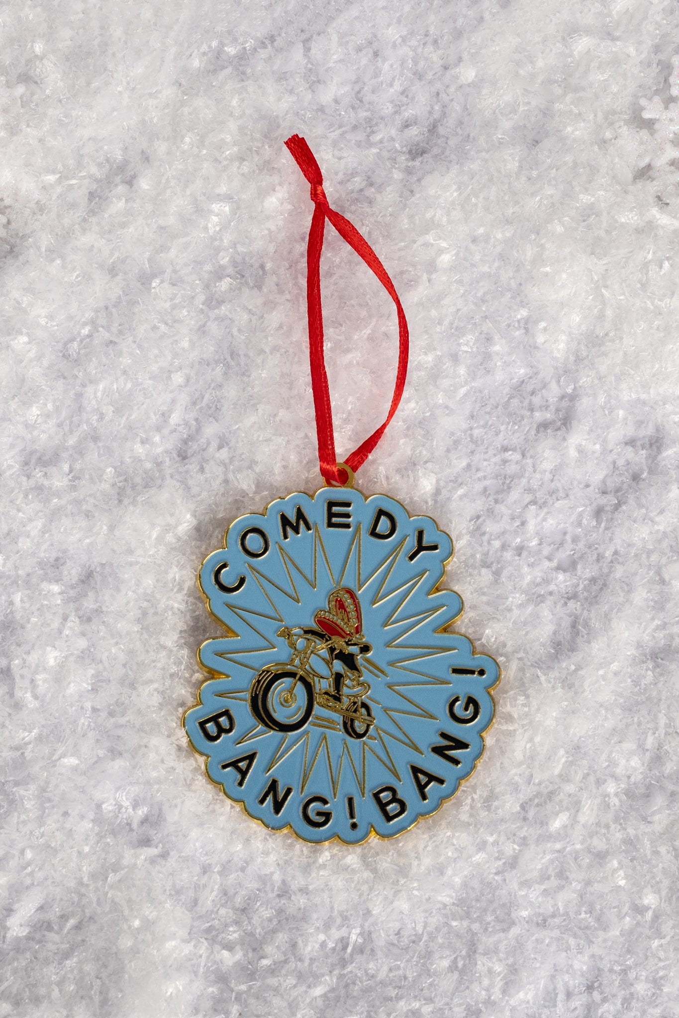 A blue enamel ornament featuring the phrase ‘COMEDY BANG! BANG!’ encircling an illustration of a biker wearing a red helmet, shown on snow-like textured ground.