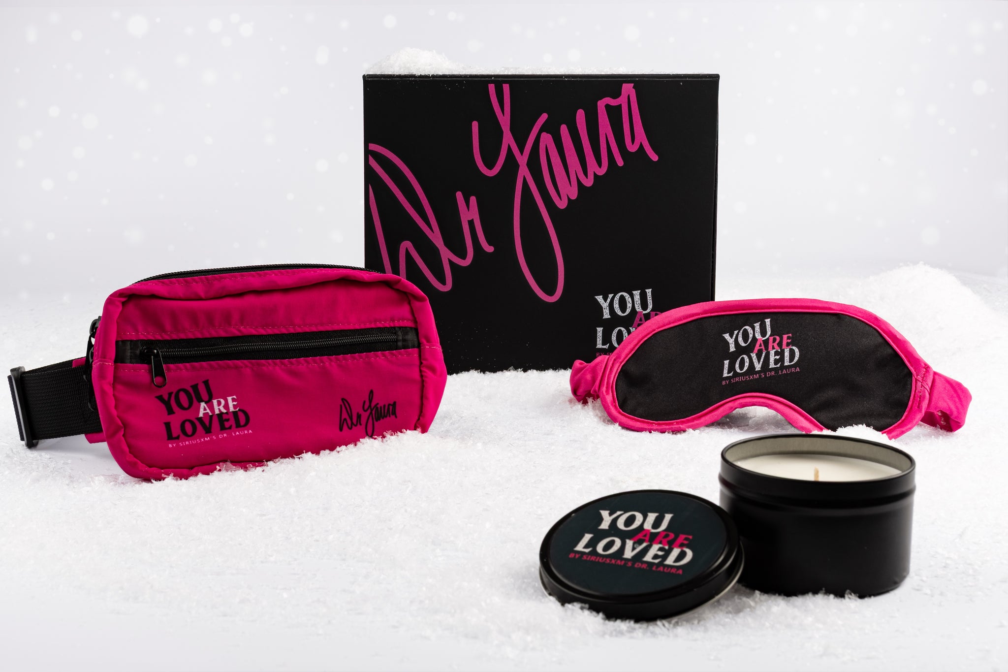 A snowy holiday flat lay of SiriusXM’s You Are Loved by Dr. Laura gift set, featuring a bright pink hip pack with ‘YOU ARE LOVED by SiriusXM’s Dr. Laura’ and Dr. Laura’s signature in black, a black and pink sleep mask displaying the same phrase, and a black candle tin with its lid showing the same printed text. Behind the items, a black gift box with Dr. Laura’s large pink signature and the ‘YOU ARE LOVED by SiriusXM’s Dr. Laura’ logo in white and pink is positioned as the backdrop.