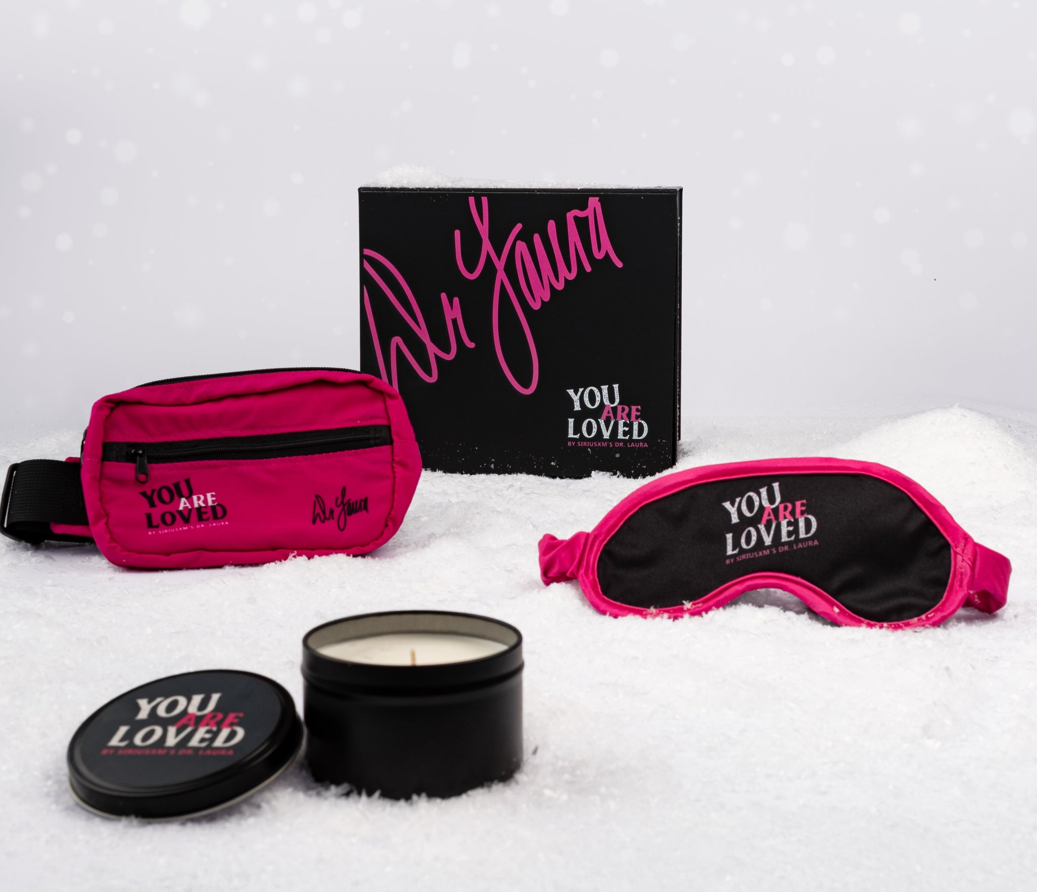 A holiday-themed flat lay of SiriusXM’s You Are Loved by Dr. Laura gift set displayed on white snow. The set includes a bright pink hip pack printed with the text ‘YOU ARE LOVED by SiriusXM’s Dr. Laura’ and Dr. Laura’s signature in black, a black and pink sleep mask featuring the same ‘YOU ARE LOVED’ text, and a small black candle tin with a matching printed lid. Behind them sits a matte black gift box with Dr. Laura’s signature in large pink script across the top and the words ‘YOU ARE LOVED by SiriusXM’s 