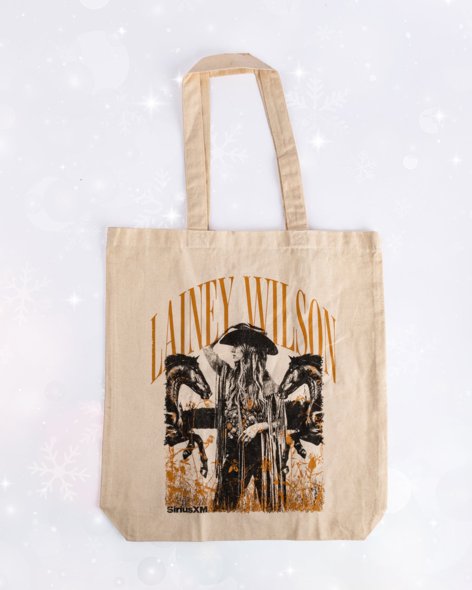 A tan canvas tote bag printed with an image of Lainey Wilson wearing a hat and fringe jacket beside two horses. Above her, the text reads ‘LAINEY WILSON,’ with the ‘SiriusXM’ logo printed below.