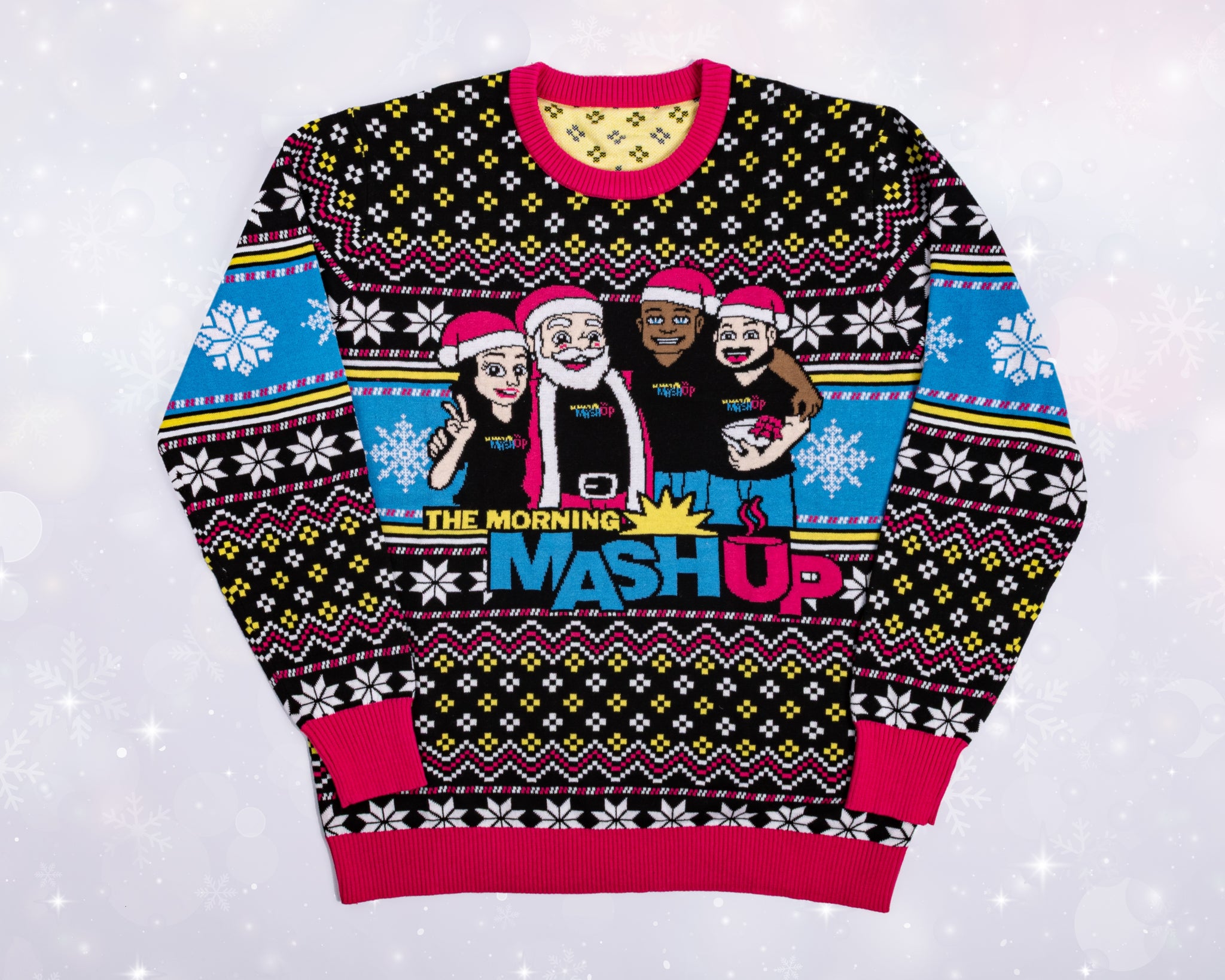A flat lay of a colorful holiday sweater featuring a cartoon design of The Morning Mash Up team wearing Santa hats. The sweater has a black, blue, and yellow geometric snowflake pattern with pink trim at the collar, cuffs, and hem. The center graphic shows three hosts and Santa posing together above the text ‘THE MORNING MASH UP’ in bold yellow, blue, and pink letters.