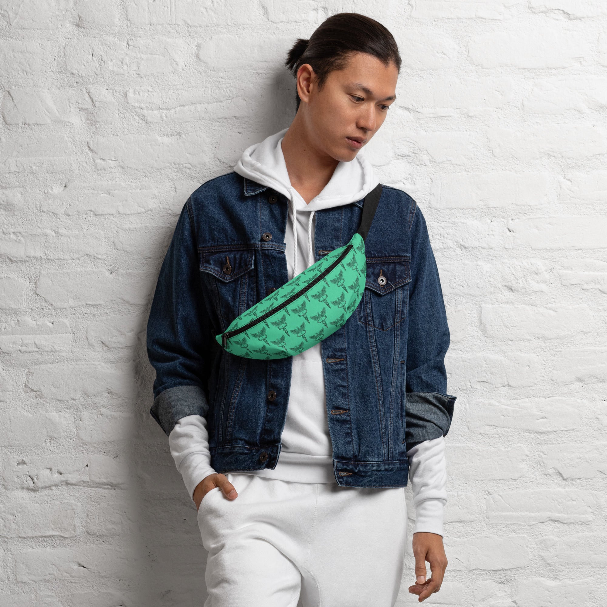 A person with long dark hair tied back is leaning against a white brick wall while wearing a bright green fanny pack with a repeating black pattern of winged staffs entwined by two serpents — a symbol commonly associated with medicine and healthcare. The fanny pack is worn crossbody over a white hoodie layered under a denim jacket, and it features a black adjustable strap.
