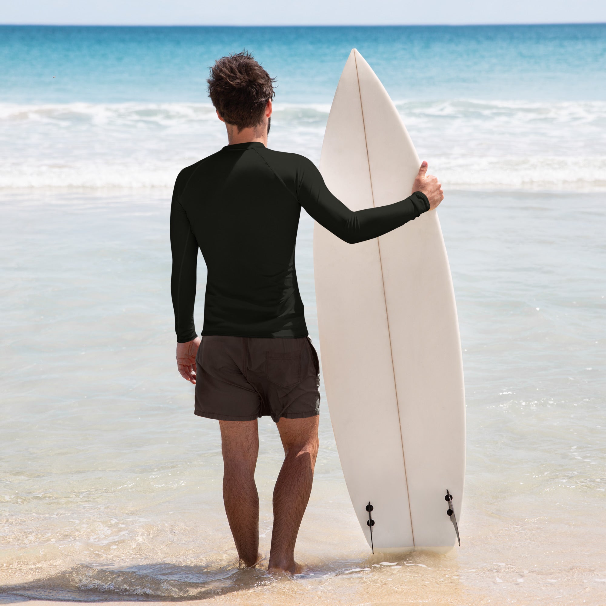 A man stands at the shoreline with a white surfboard under his arm, wearing a plain black long-sleeve rash guard and black swim trunks, facing the ocean.