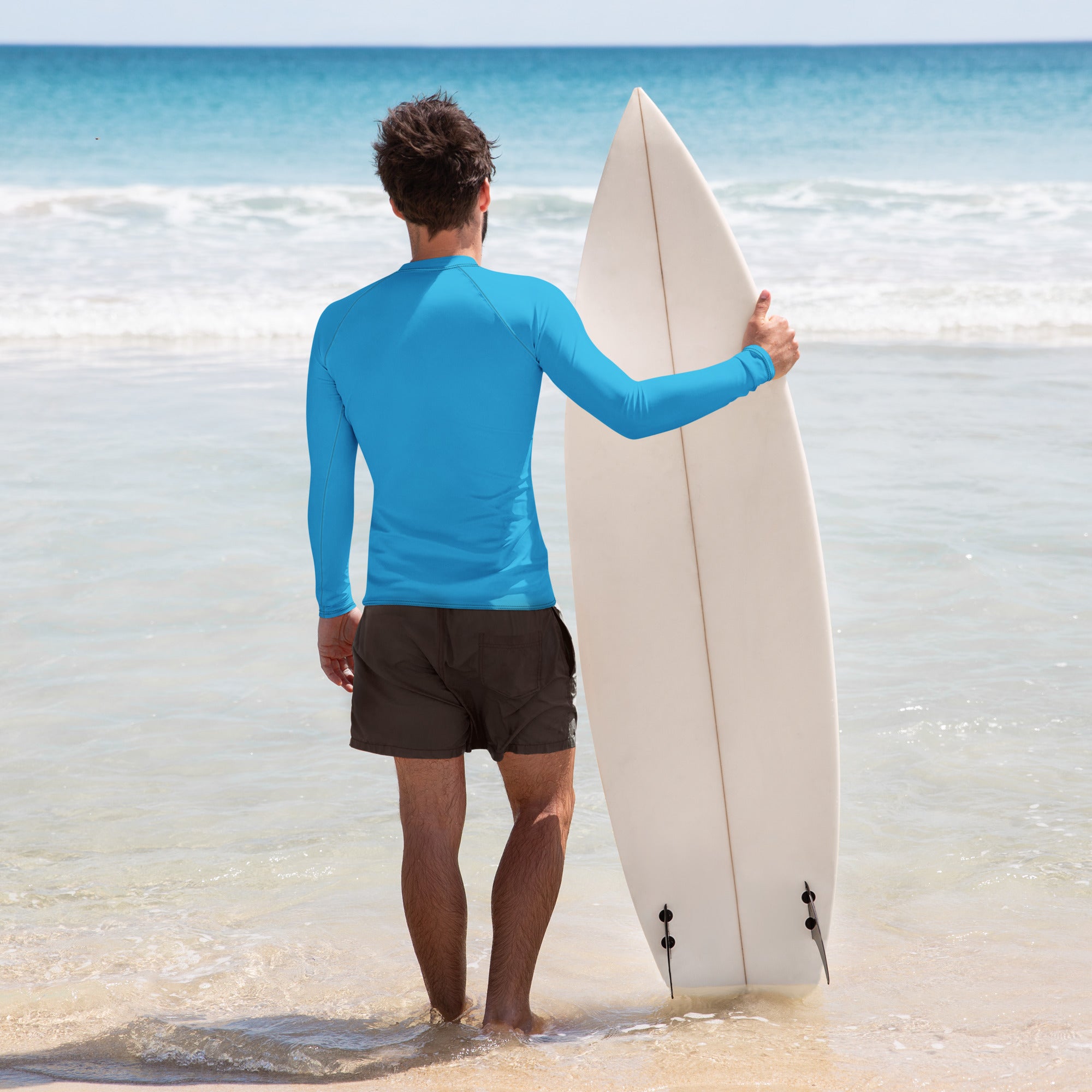 A man stands in ankle-deep ocean water holding a white surfboard, wearing a bright blue long-sleeve rash guard and black swim trunks. The back of the shirt is plain with no visible design.