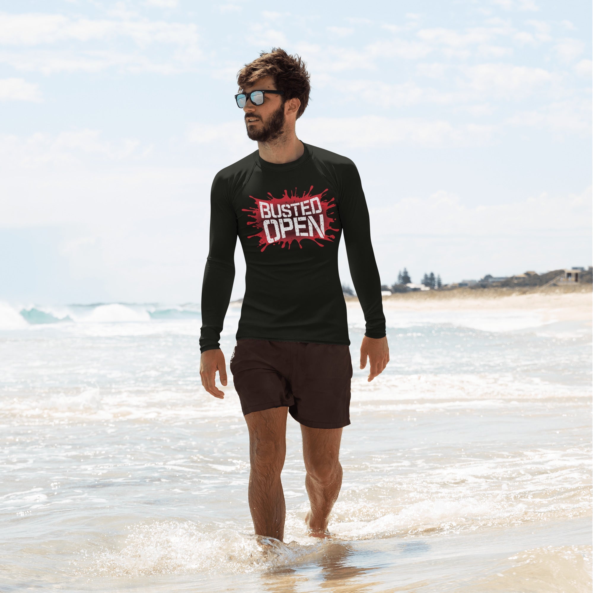 A man walks in shallow water at the beach wearing a black long-sleeve rash guard featuring a bold red graphic that reads 'BUSTED OPEN' in white, with a red splatter background. He pairs it with black swim trunks and sunglasses.
