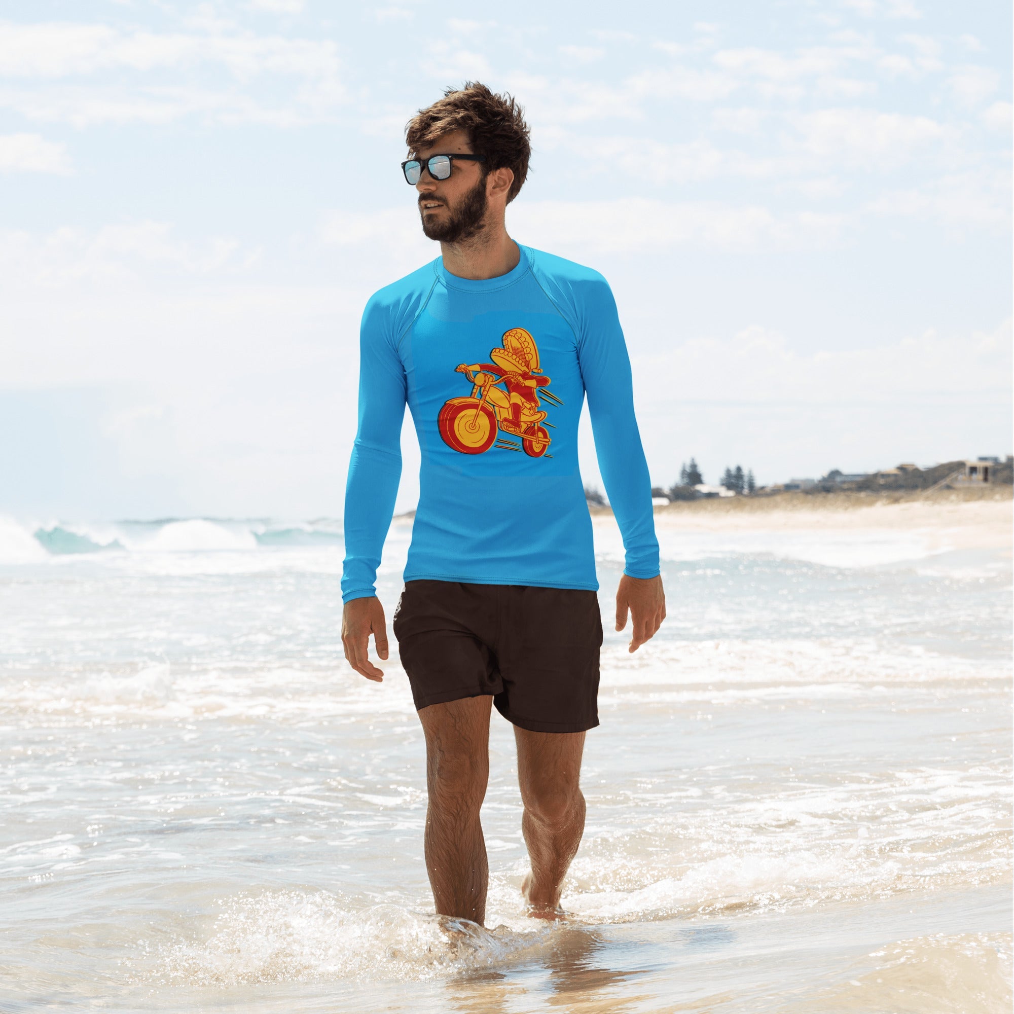 A man walks along the shoreline wearing a bright blue long-sleeve rash guard with a bold graphic of Comedy Bang Bang's classic red and yellow Motormouth character, riding a motorcycle. He pairs it with black swim trunks and sunglasses.