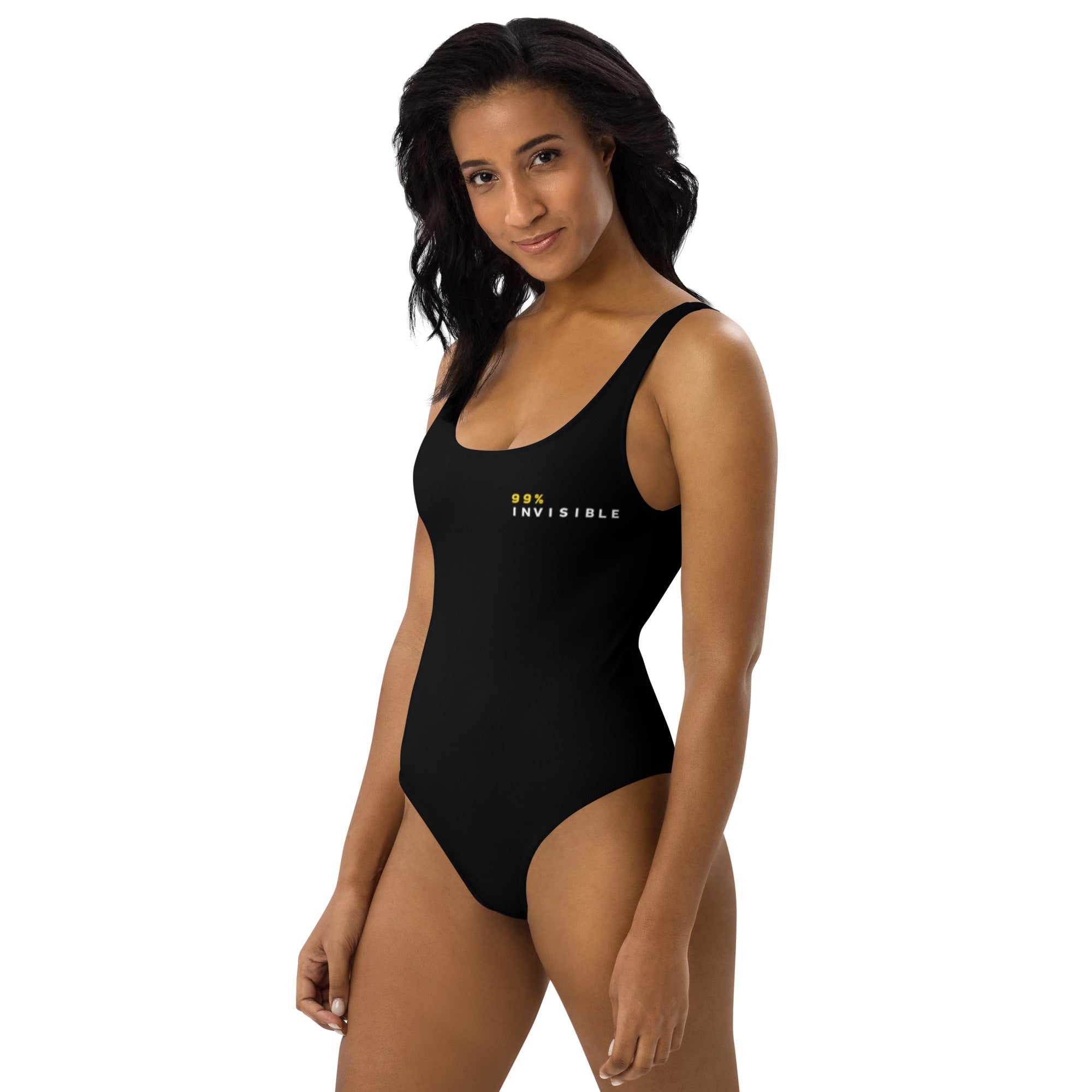 A woman wearing a black one-piece swimsuit featuring the text '99% INVISIBLE' in yellow and white on the left chest, smiling slightly while standing against a white background.