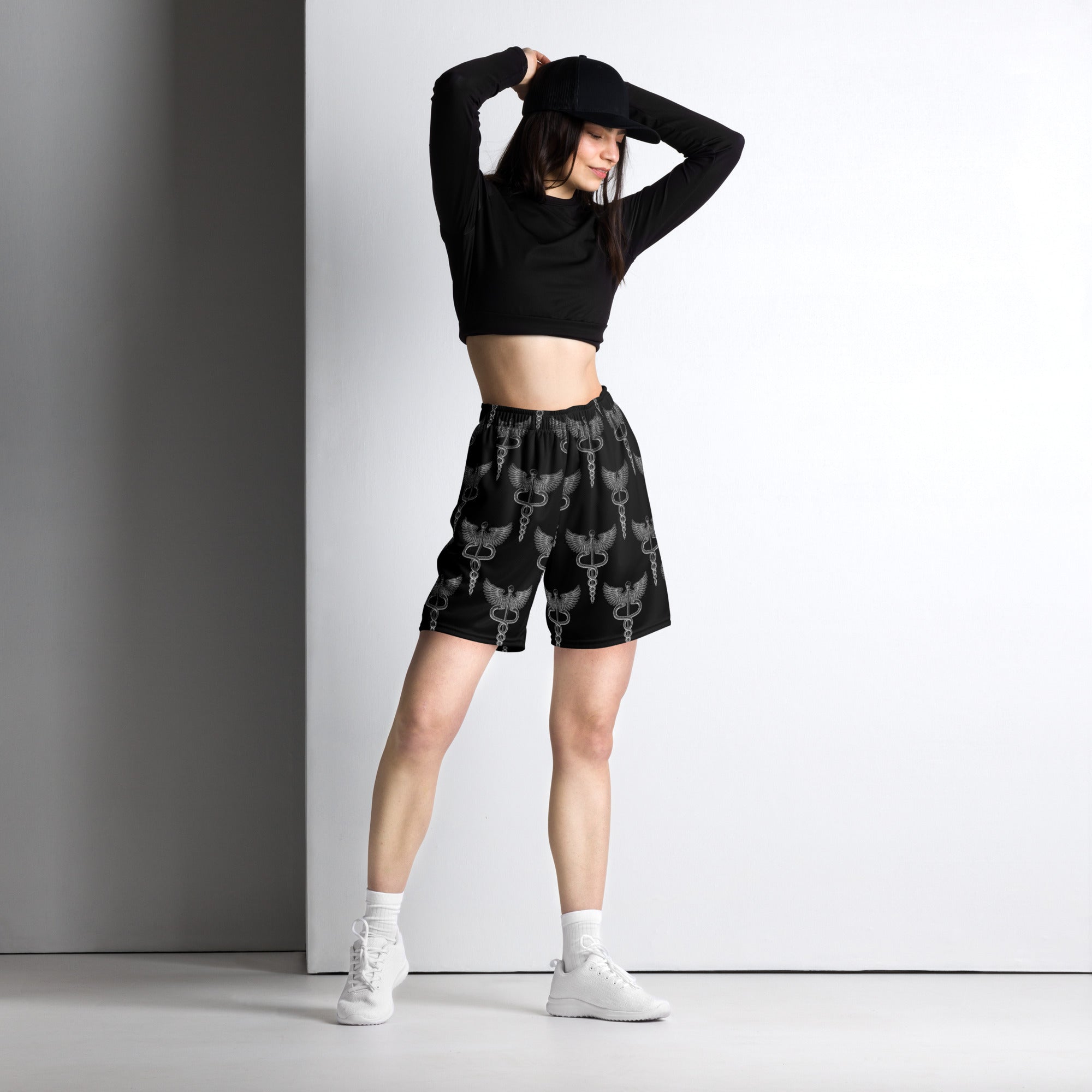 Woman poses with arms raised, wearing black mesh shorts printed with a repeating caduceus symbol—a winged staff entwined by two snakes. Paired with a black long-sleeve crop top and white sneakers.