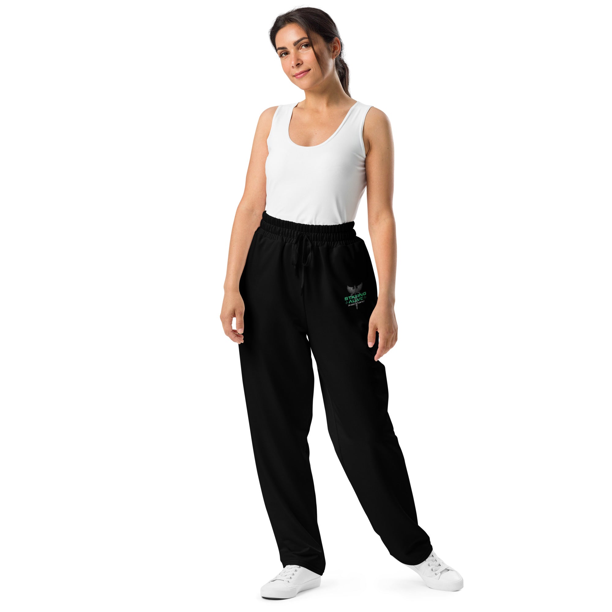 A  woman modeling black wide-leg joggers with a relaxed fit and the 'STAYING ALIVE' caduceus graphic on the left thigh, standing in white sneakers and a white tank top.