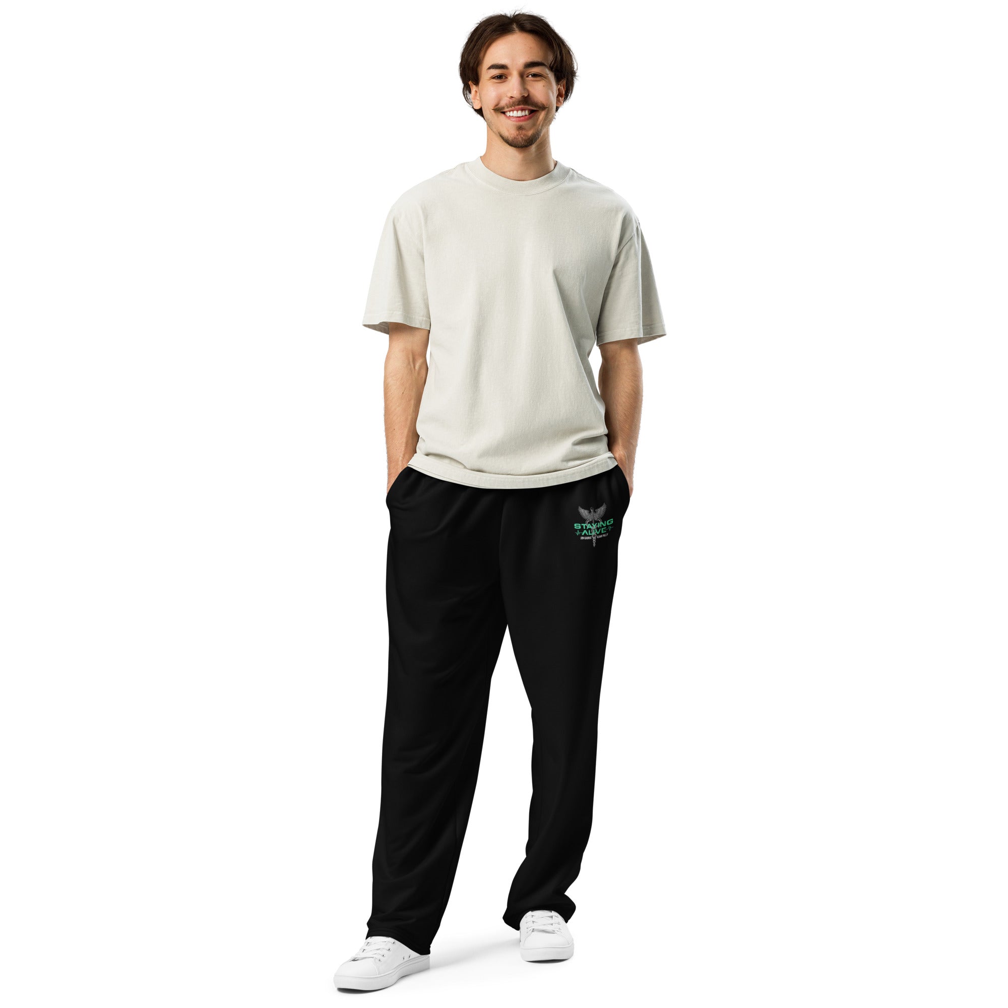 A man standing with hands in his pockets wearing black wide-leg joggers featuring a green and gray 'STAYING ALIVE' logo with a caduceus symbol—depicted as a winged staff with two snakes—on the left thigh.