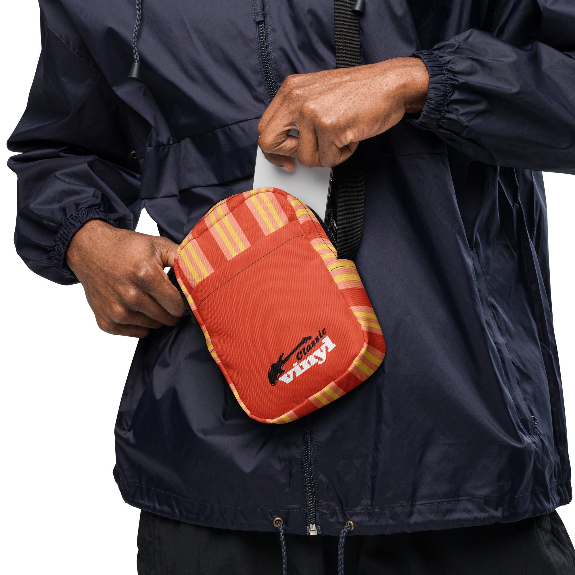 A person wearing a navy windbreaker is seen placing a phone into an orange and yellow striped crossbody bag with a black strap. The front pocket of the bag features the 'Classic vinyl' logo with a black guitar silhouette and white and red text.