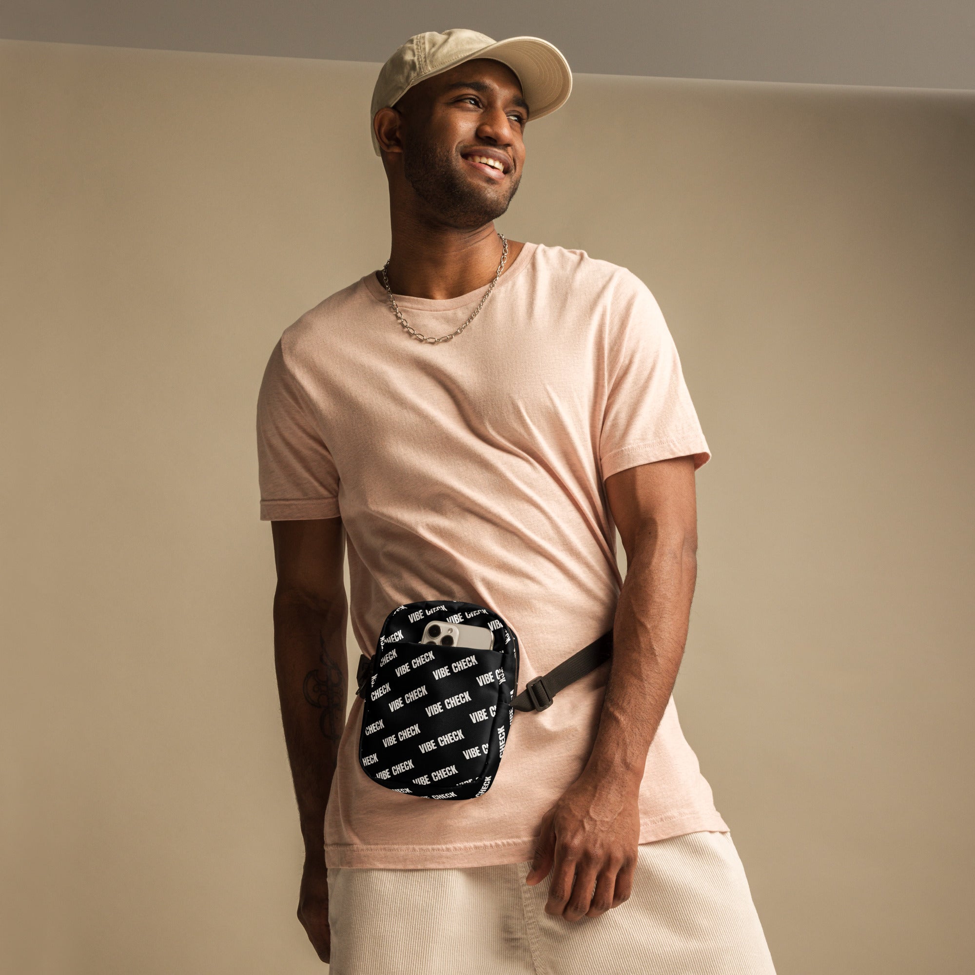 A man poses in a neutral studio wearing a pale pink t-shirt, cream shorts, and a beige cap. He has a black crossbody bag with a white repeating 'VIBE CHECK' print strapped around his waist.
