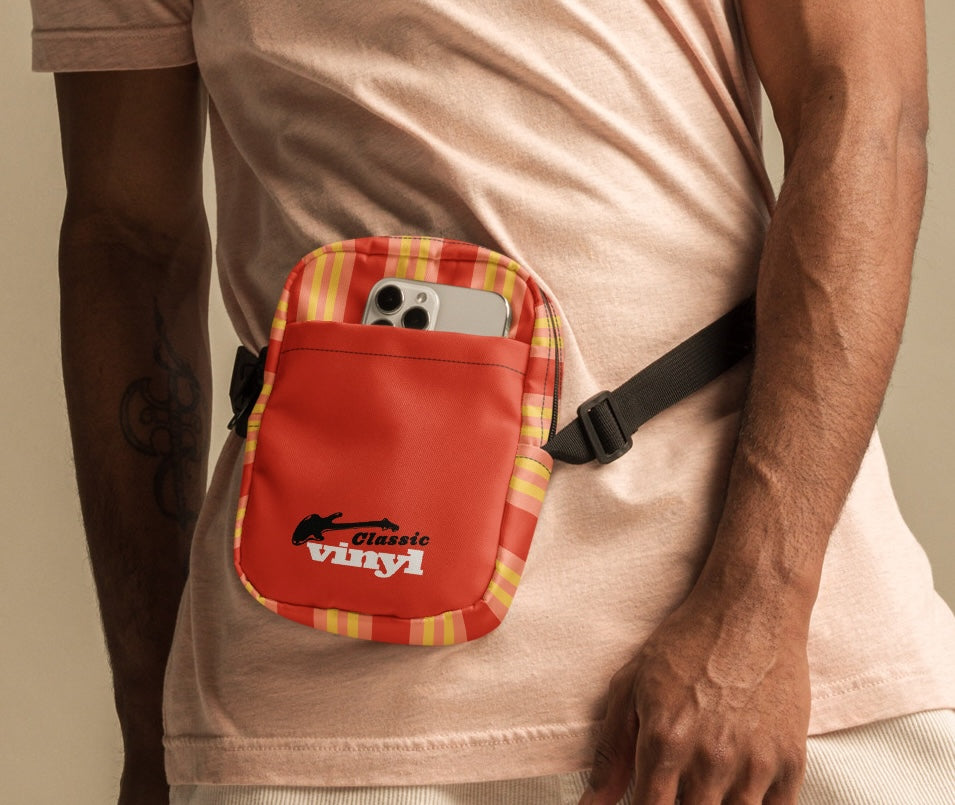 A person wearing a light pink T-shirt and cream shorts is shown with an orange and yellow striped 'Classic vinyl' bag worn around the waist. A phone is visible in the front pocket above the black strap.