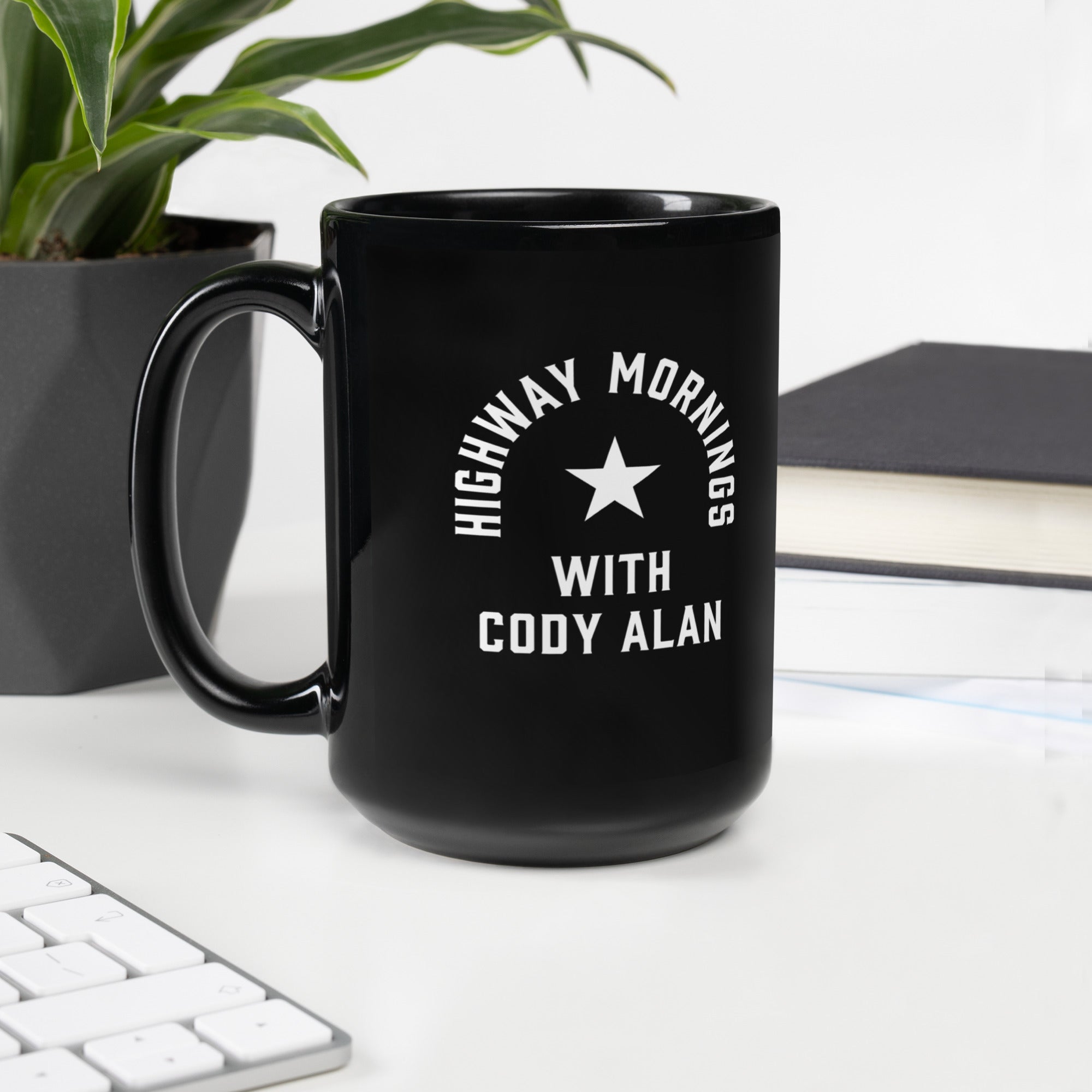 A black glossy mug with the phrase 'HIGHWAY MORNINGS WITH CODY ALAN' and a white star in the center, placed on a white desktop near a keyboard, book, and potted plant.