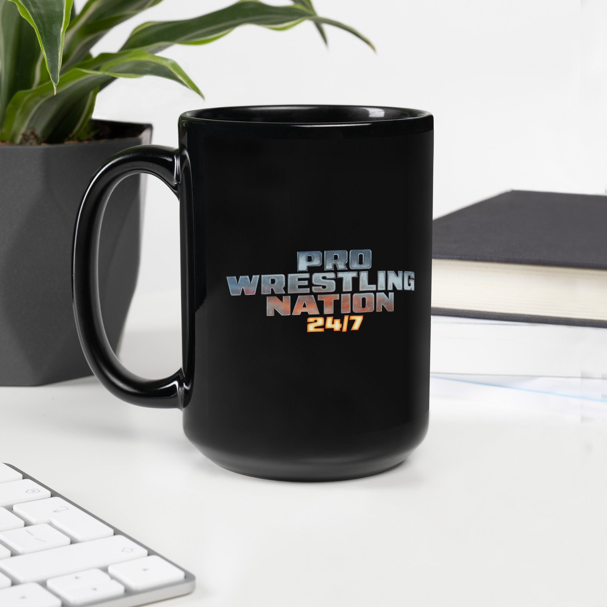Black glossy ceramic mug featuring the 'Pro Wrestling Nation 24/7' logo in metallic gradient lettering, placed on a white desk beside a plant and a closed book.