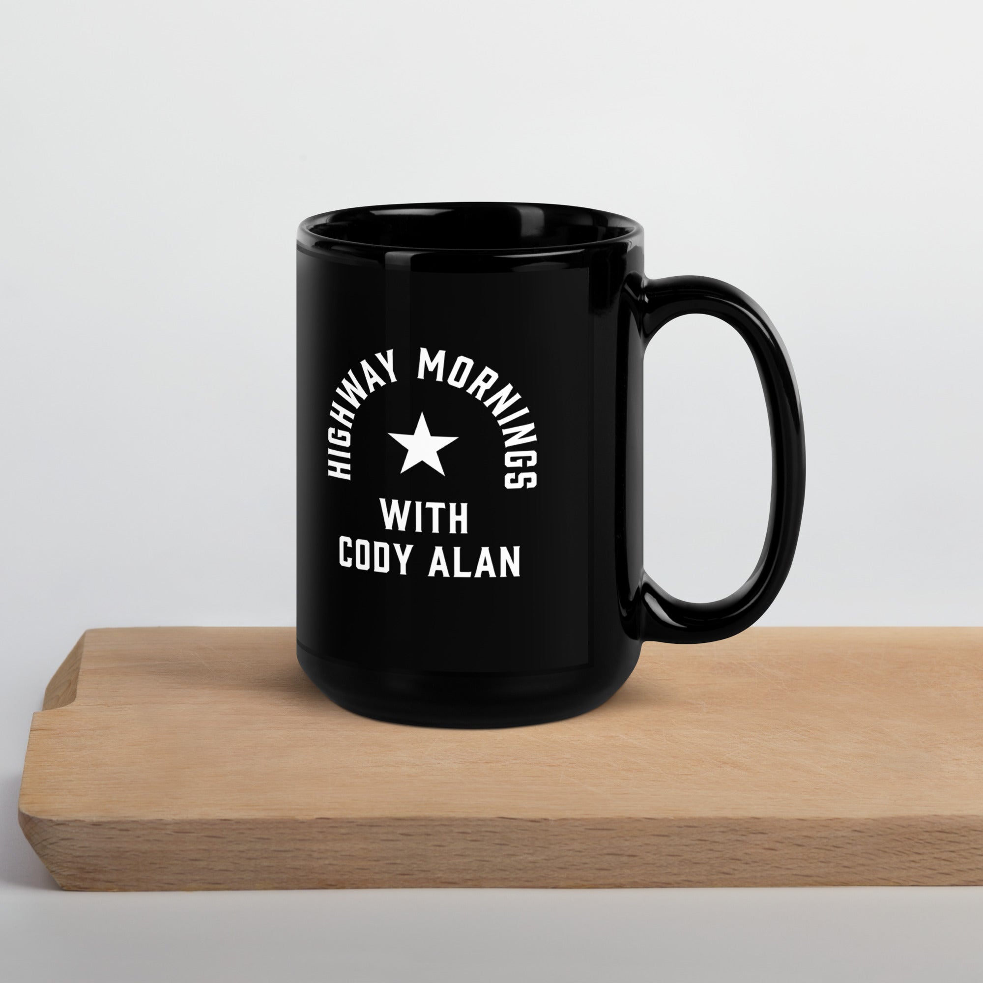 A black glossy ceramic mug featuring the text 'HIGHWAY MORNINGS WITH CODY ALAN' in white and a central star, set on a light wooden serving tray against a neutral background.