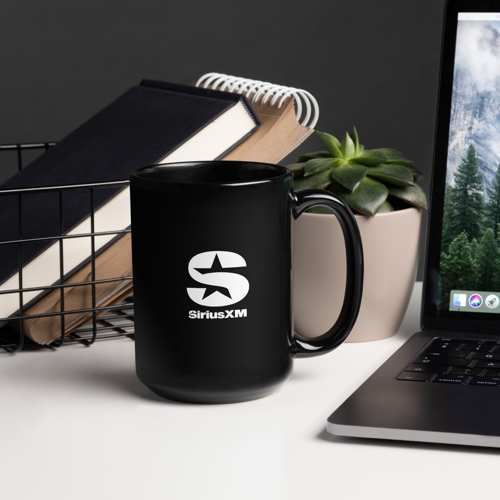 Black ceramic mug with white 'SiriusXM' star logo on front, styled on a modern desk with books, a succulent plant, and a laptop in the background.