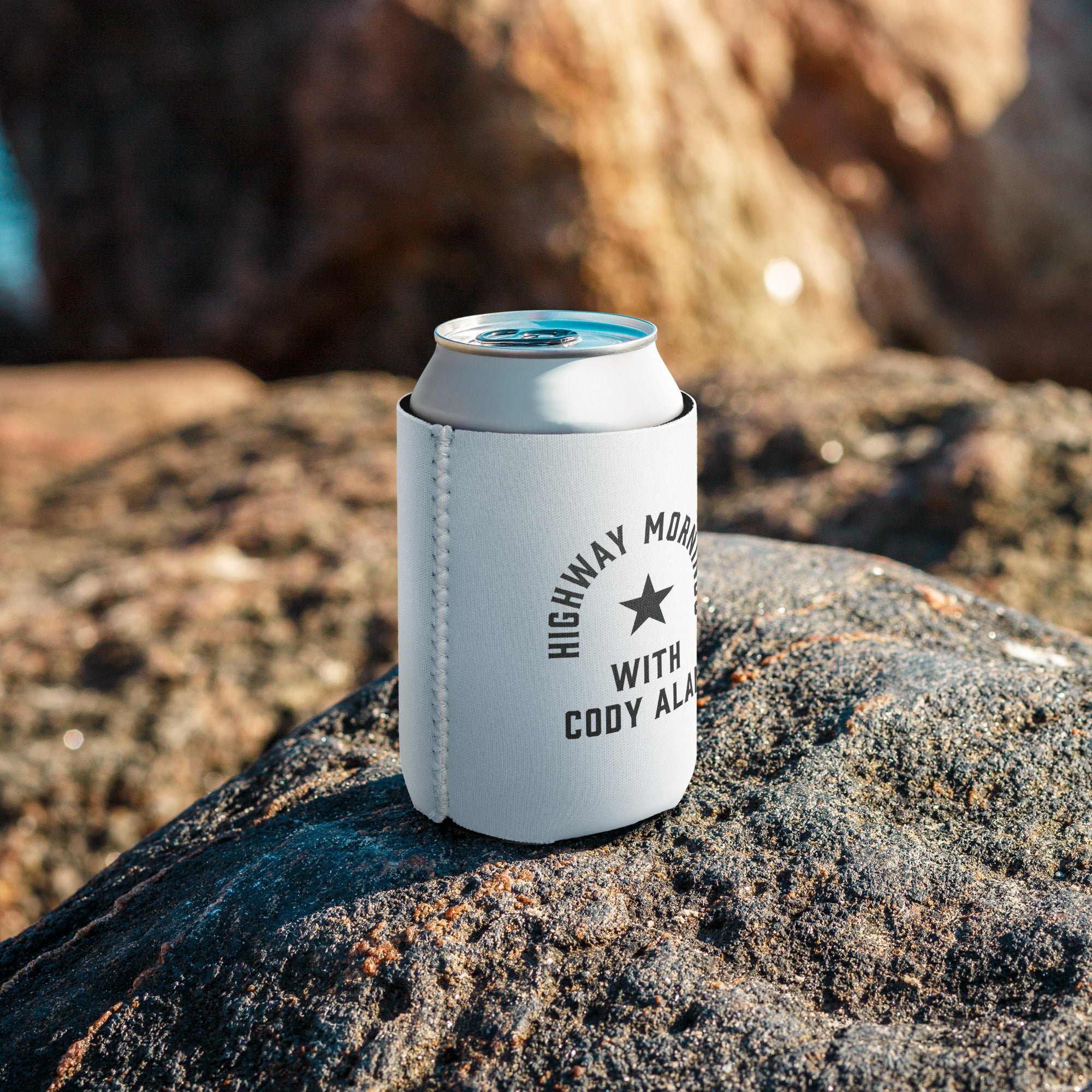 A white can cooler with the 'HIGHWAY MORNINGS WITH CODY ALAN' design resting on a rock surface, wrapped around a silver can, with blurred natural surroundings in the background.