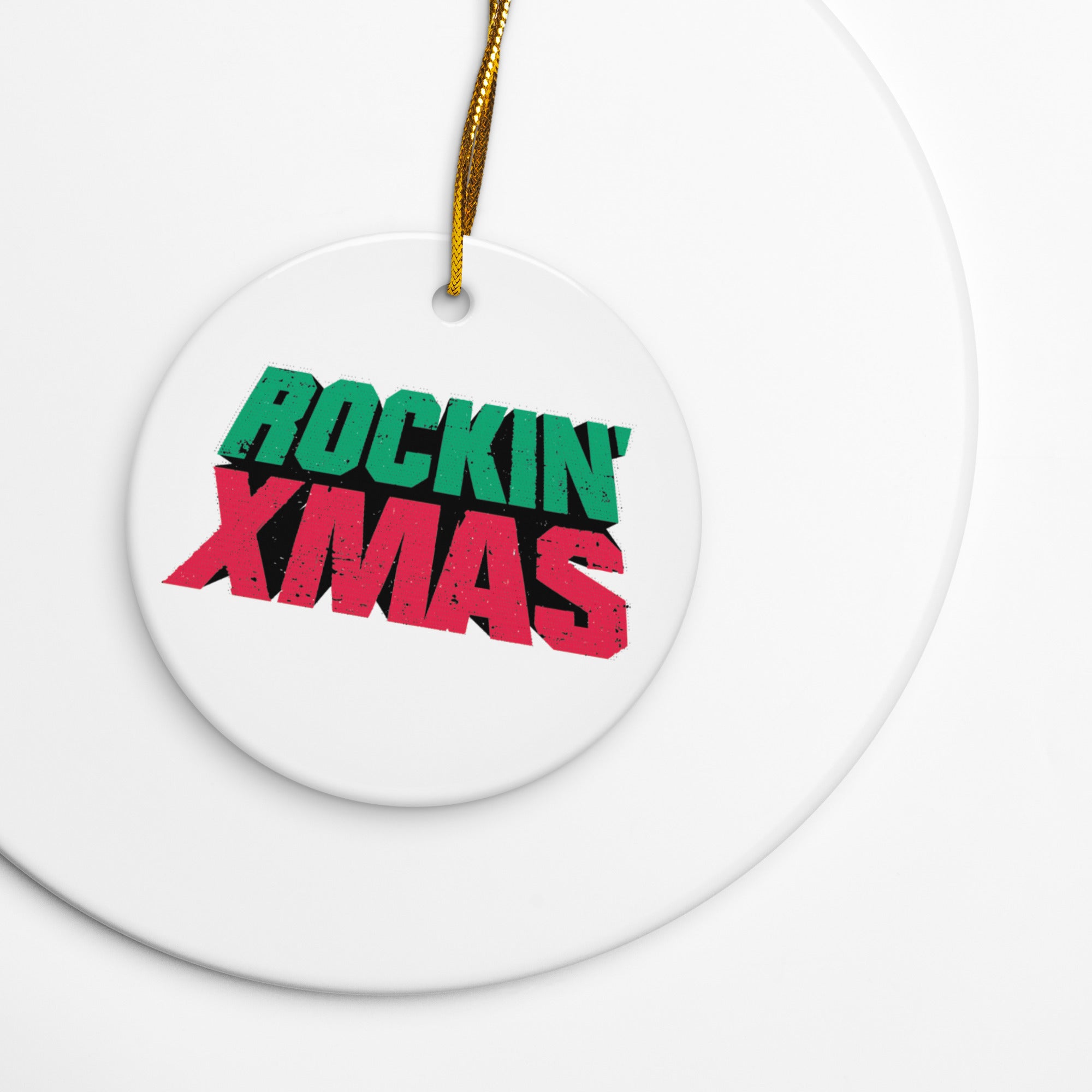 Close-up of a white round ceramic ornament with a gold hanging string, printed with the words ‘ROCKIN’ XMAS’ in bold distressed green and red letters, shown resting on a white surface.
