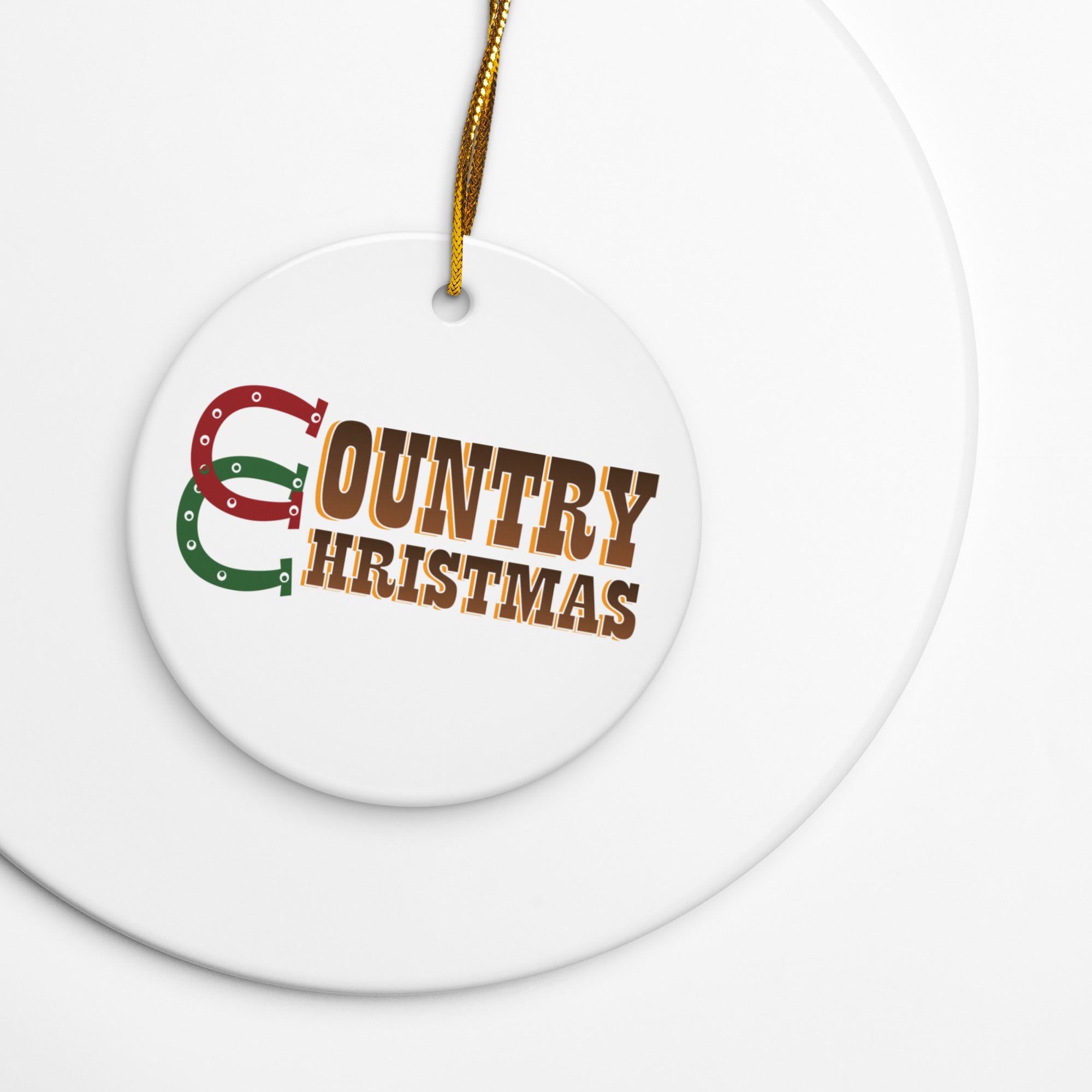Close-up of a white round ceramic ornament with a gold hanging string, printed with the words ‘COUNTRY CHRISTMAS’ in brown text and a red and green horseshoe-shaped ‘C’, resting on a white surface.