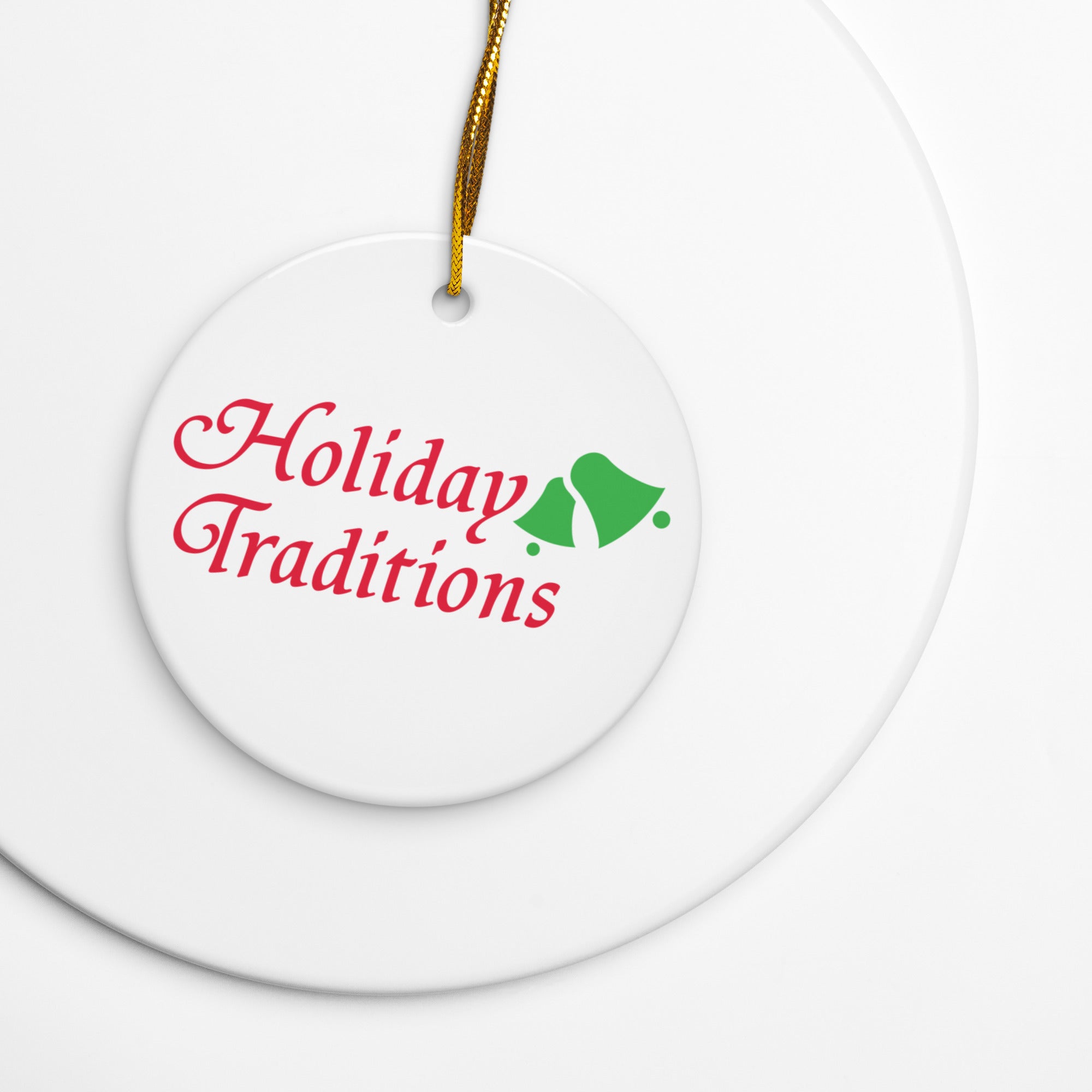 Close-up of a white round ceramic ornament with a gold hanging string, featuring the words ‘Holiday Traditions’ in red cursive text next to two green bells.
