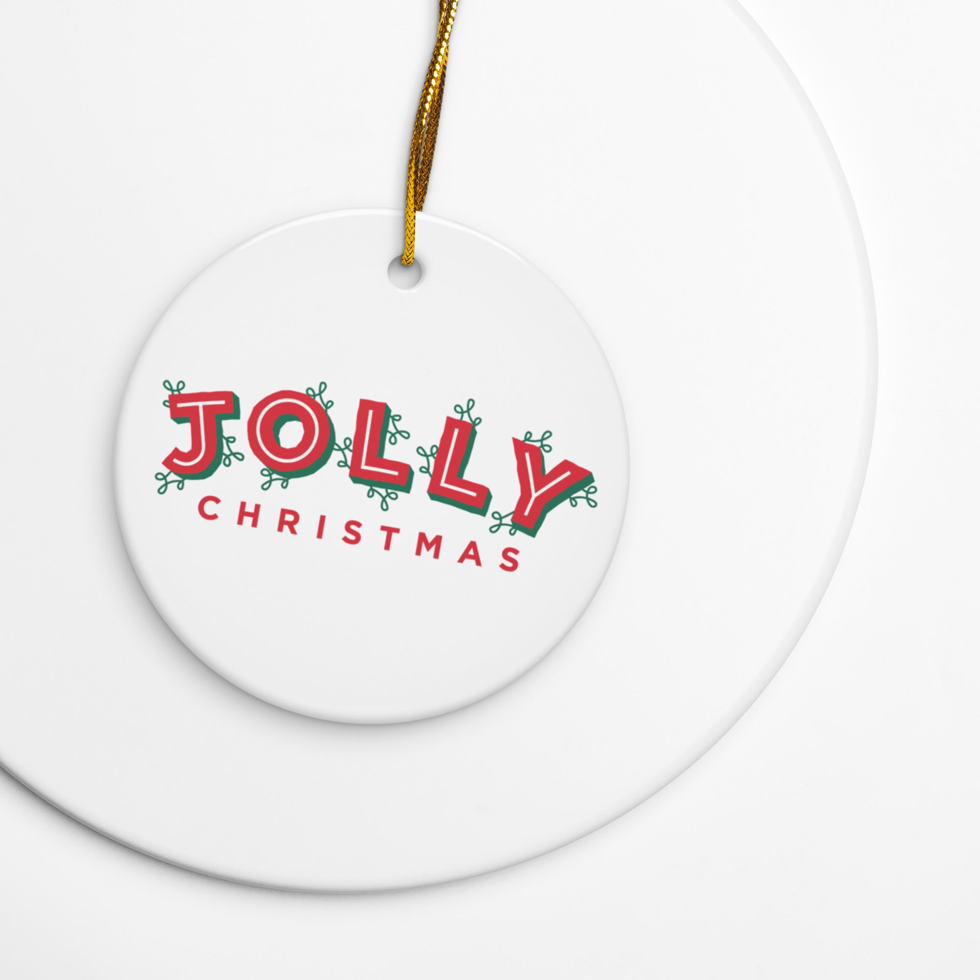 Close-up of a white ceramic ornament lying on a white surface, decorated with the phrase ‘JOLLY CHRISTMAS’ in red and green stylized letters, hanging by a gold string.