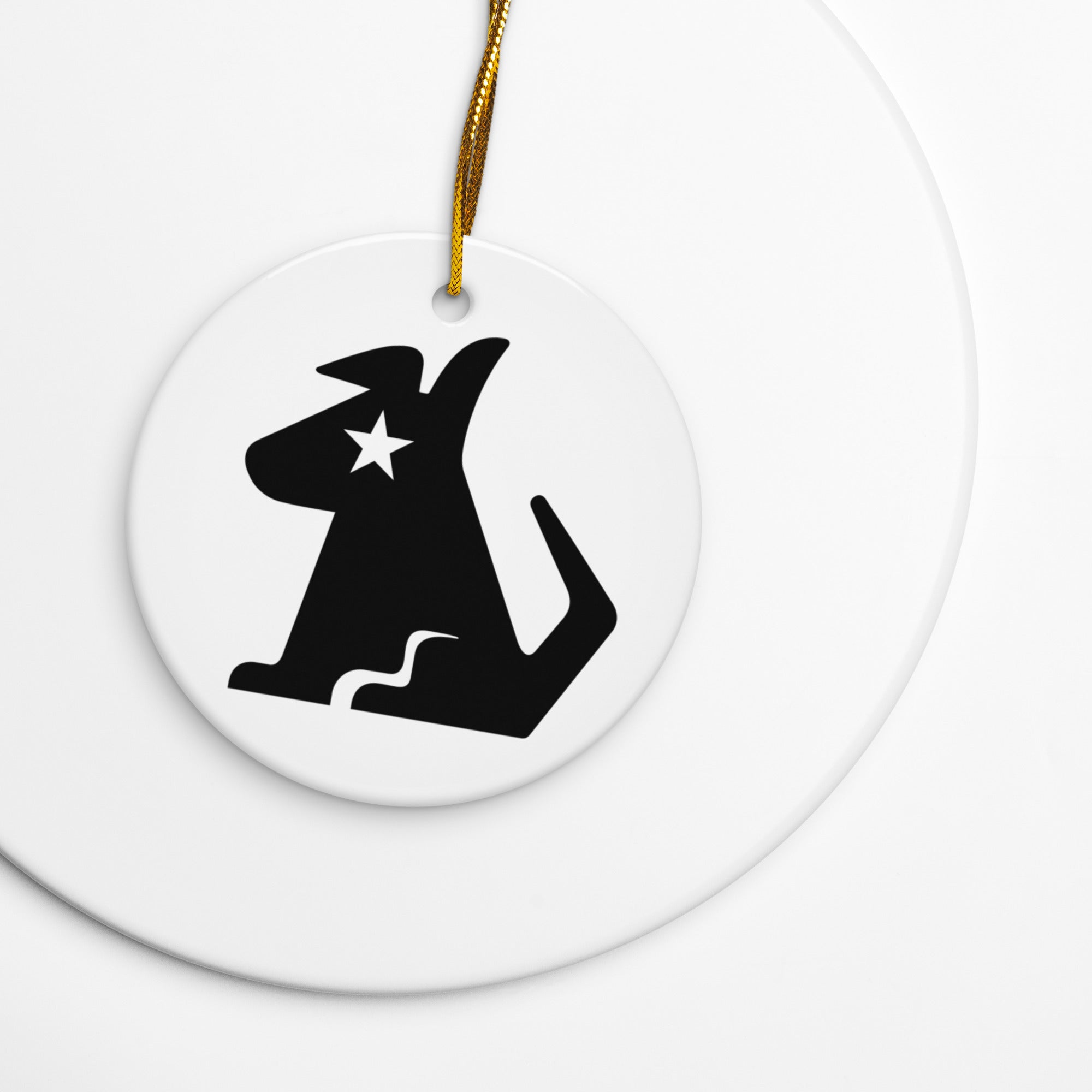 White round ceramic ornament with a gold hanging string, featuring the black silhouette of Stella Dog, with a star-shaped cutout near her eye — the signature ‘SiriusXM’ logo.