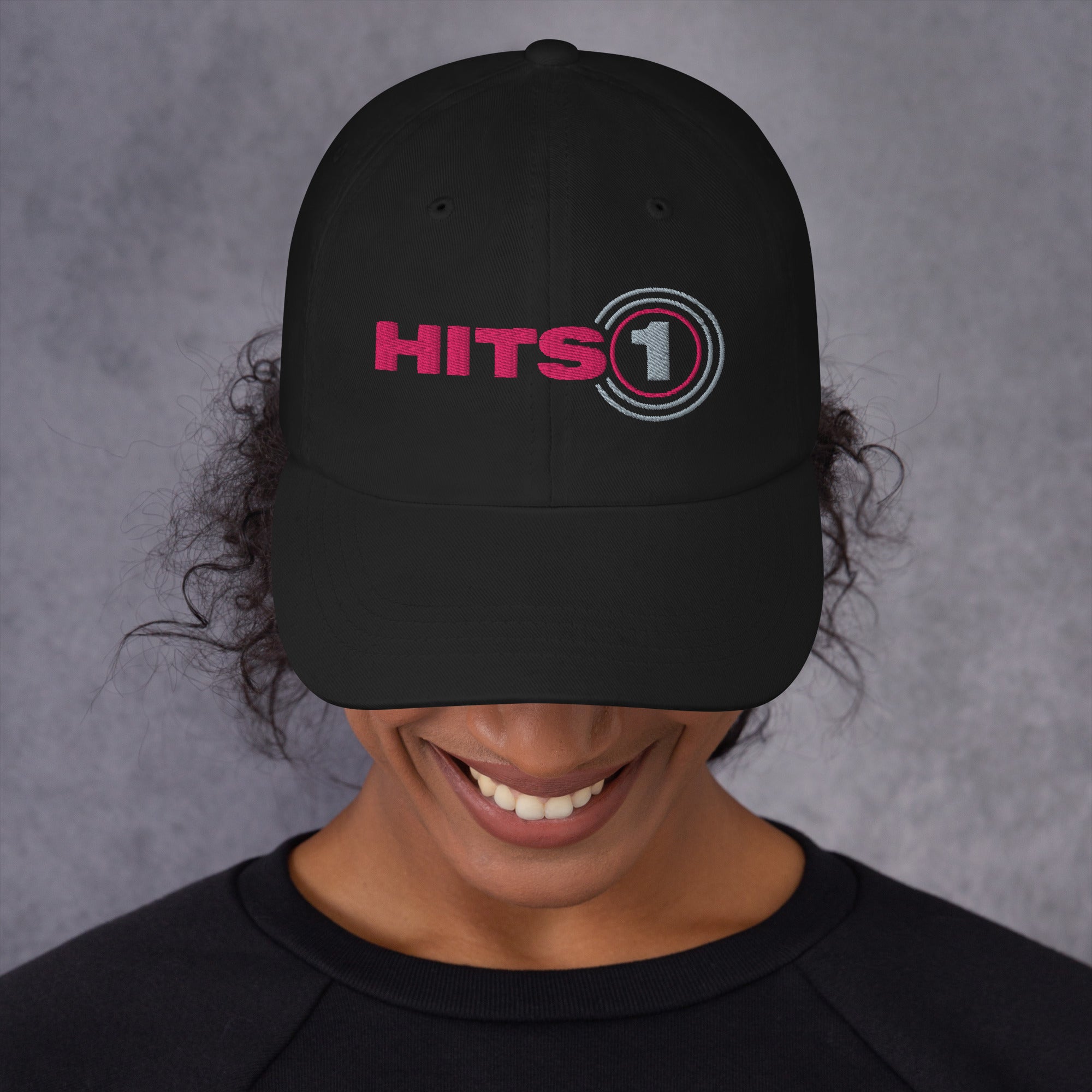 Product mockup of black hat with 'Hits 1' logo on front in pink and light grey on model.