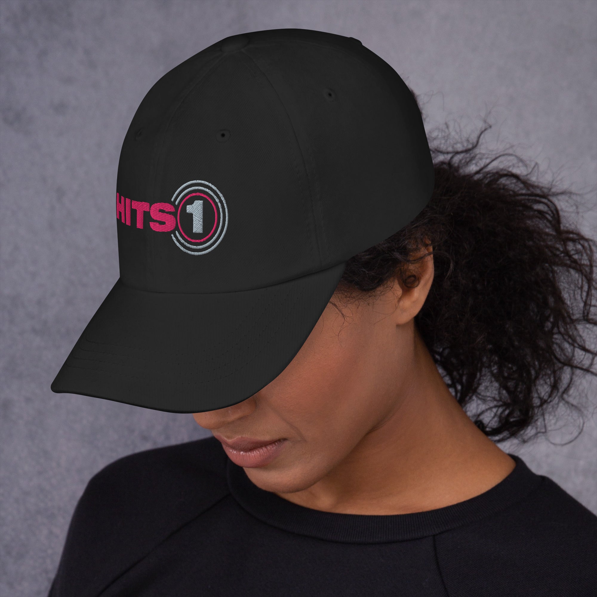 Side view of black hat with 'Hits 1' logo on front in pink and light grey on model.