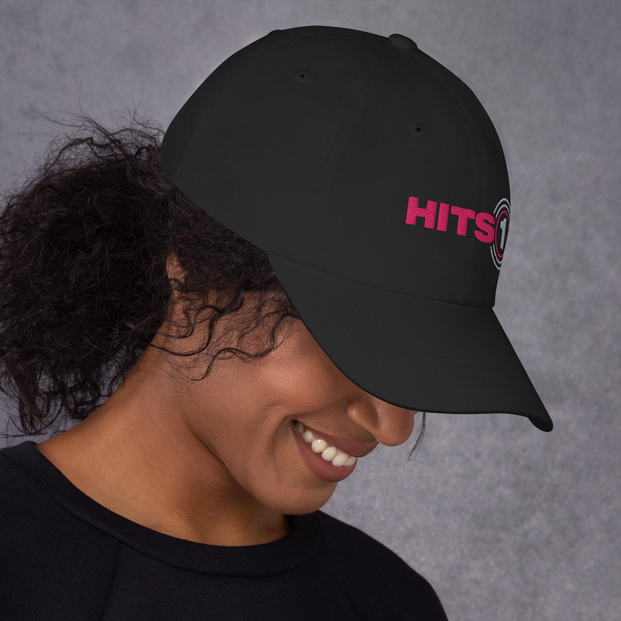 Side view of black hat with 'Hits 1' logo on front in pink and light grey on model.