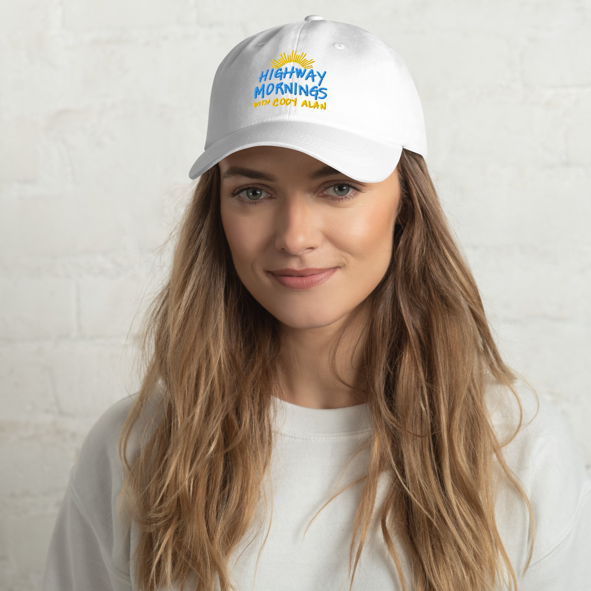 A woman wearing a white dad hat featuring the embroidered design 'HIGHWAY MORNINGS WITH CODY ALAN' in blue and yellow, with a yellow sunburst graphic above the text.