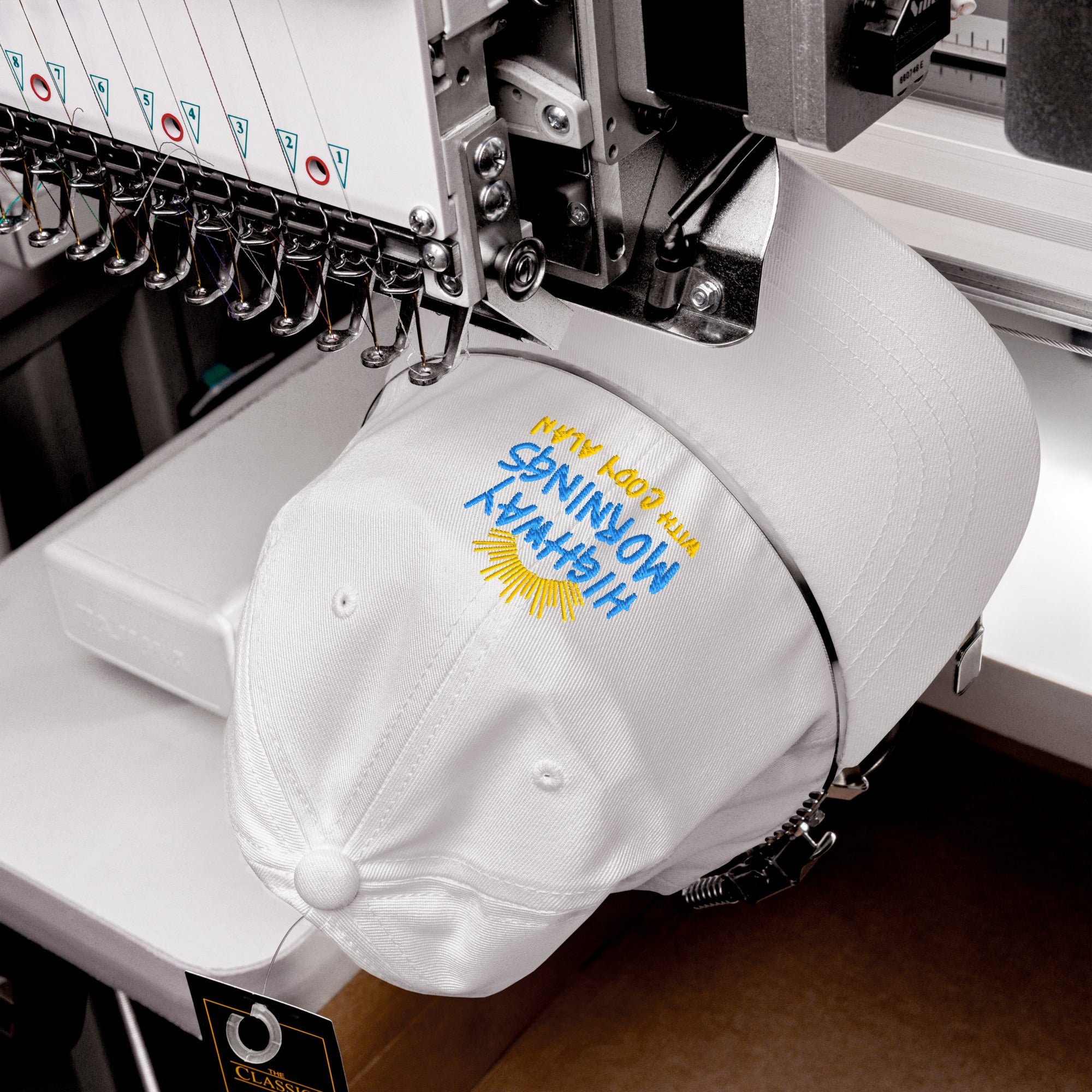 A white dad hat being embroidered on a machine, showing the design 'HIGHWAY MORNINGS WITH CODY ALAN' and sunburst being stitched in blue and yellow thread.