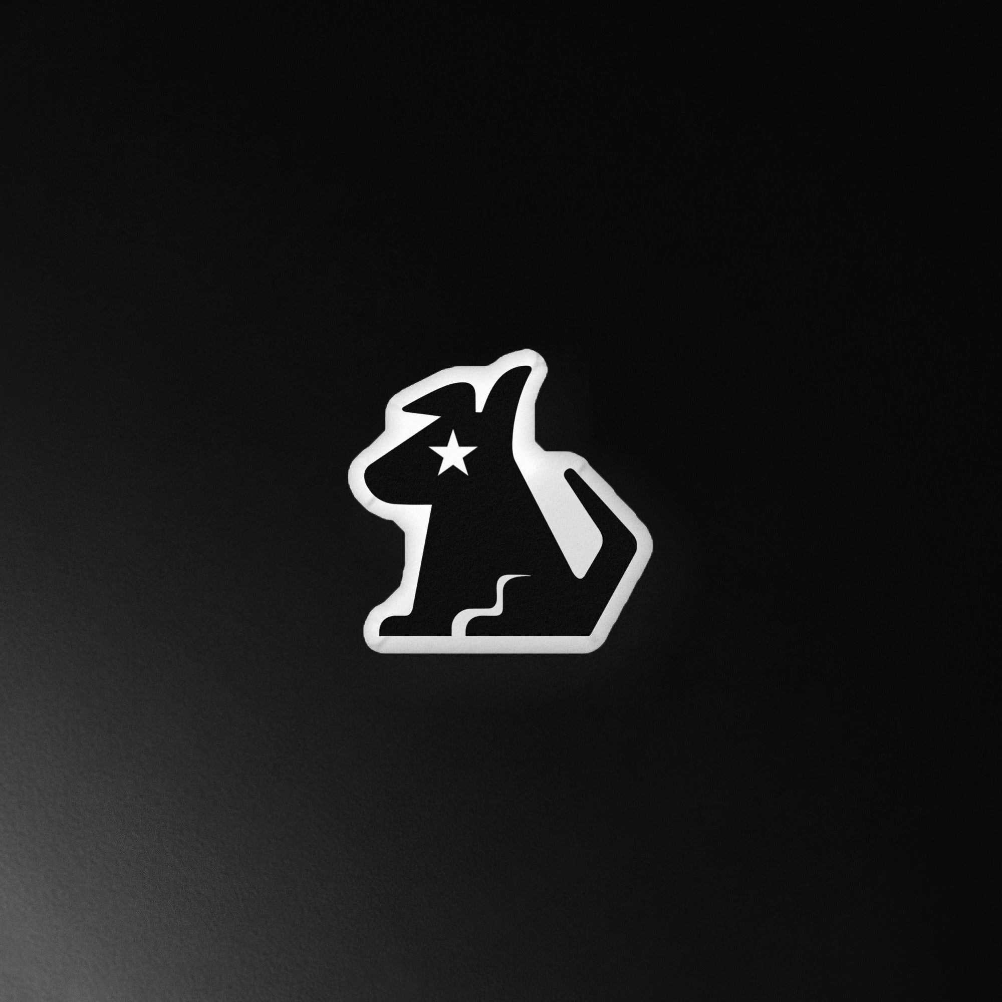 A custom-shaped black pillow in the silhouette of a dog with a pointed ear and curled tail, featuring a white star for an eye. The pillow is outlined in white and set against a dark, shadowy background for dramatic contrast.