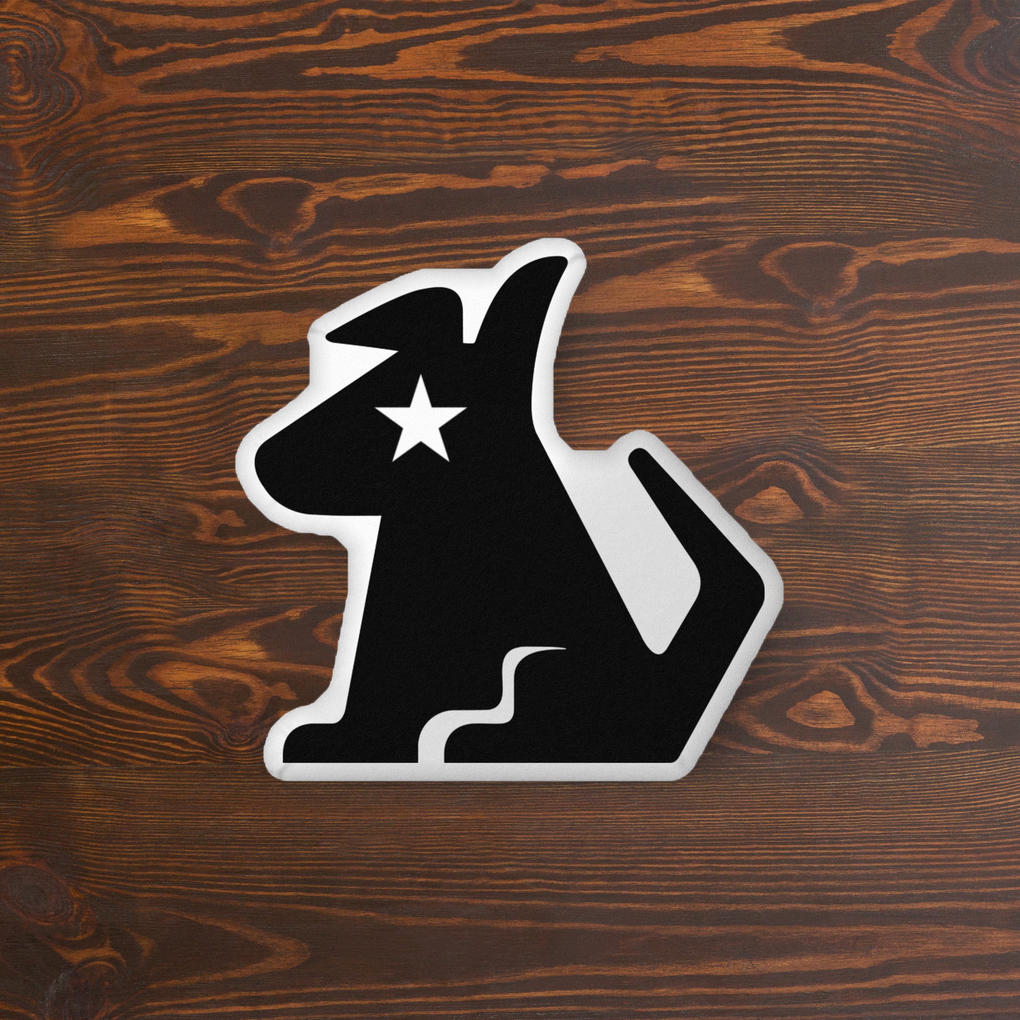 A black dog-shaped pillow with a white outline and a bold white star on its face, placed on a wooden surface with a warm, textured grain.