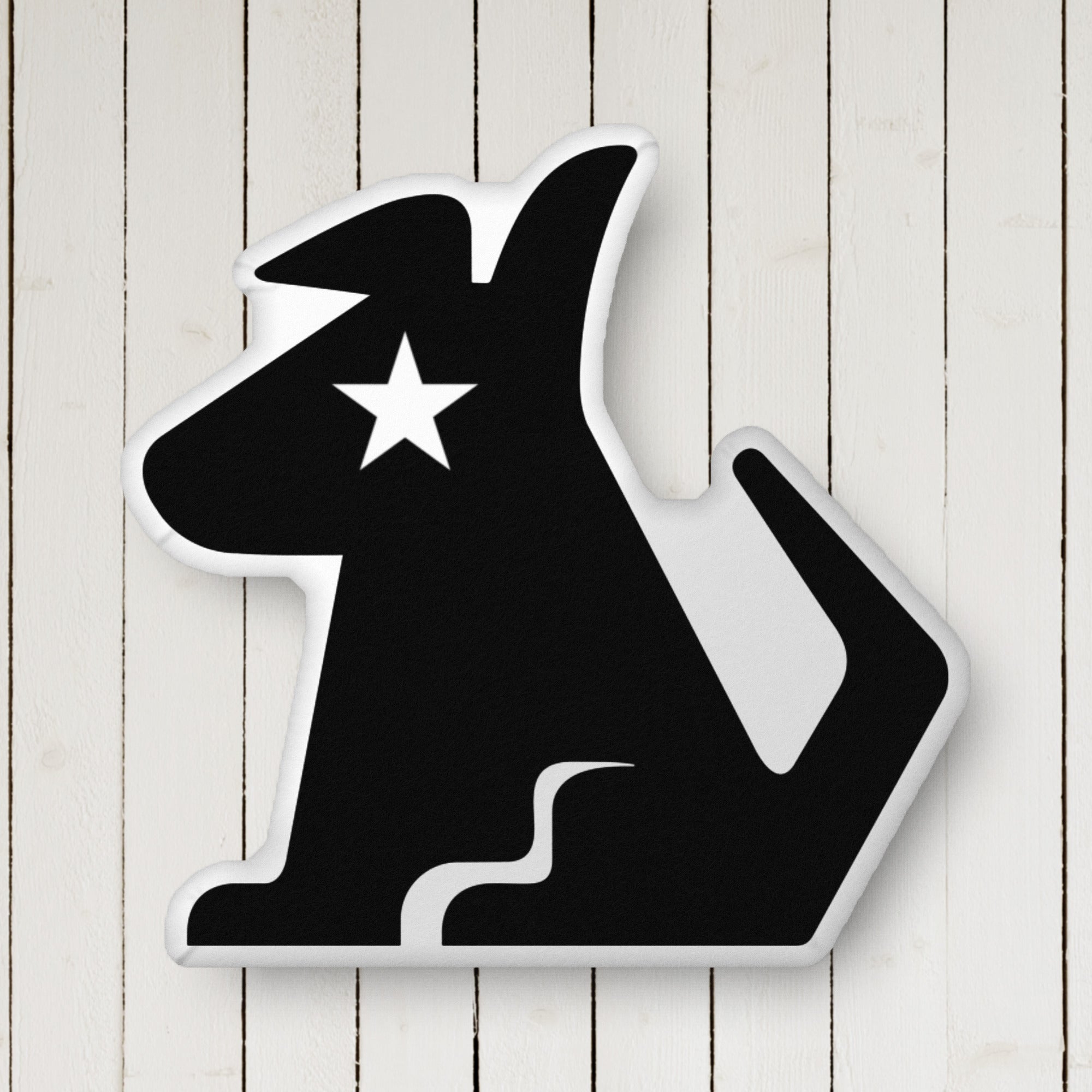 A close up of a custom-shaped black pillow in the silhouette of a dog with a pointed ear and curled tail, featuring a white star for an eye, positioned against a vertically paneled white wood backdrop.