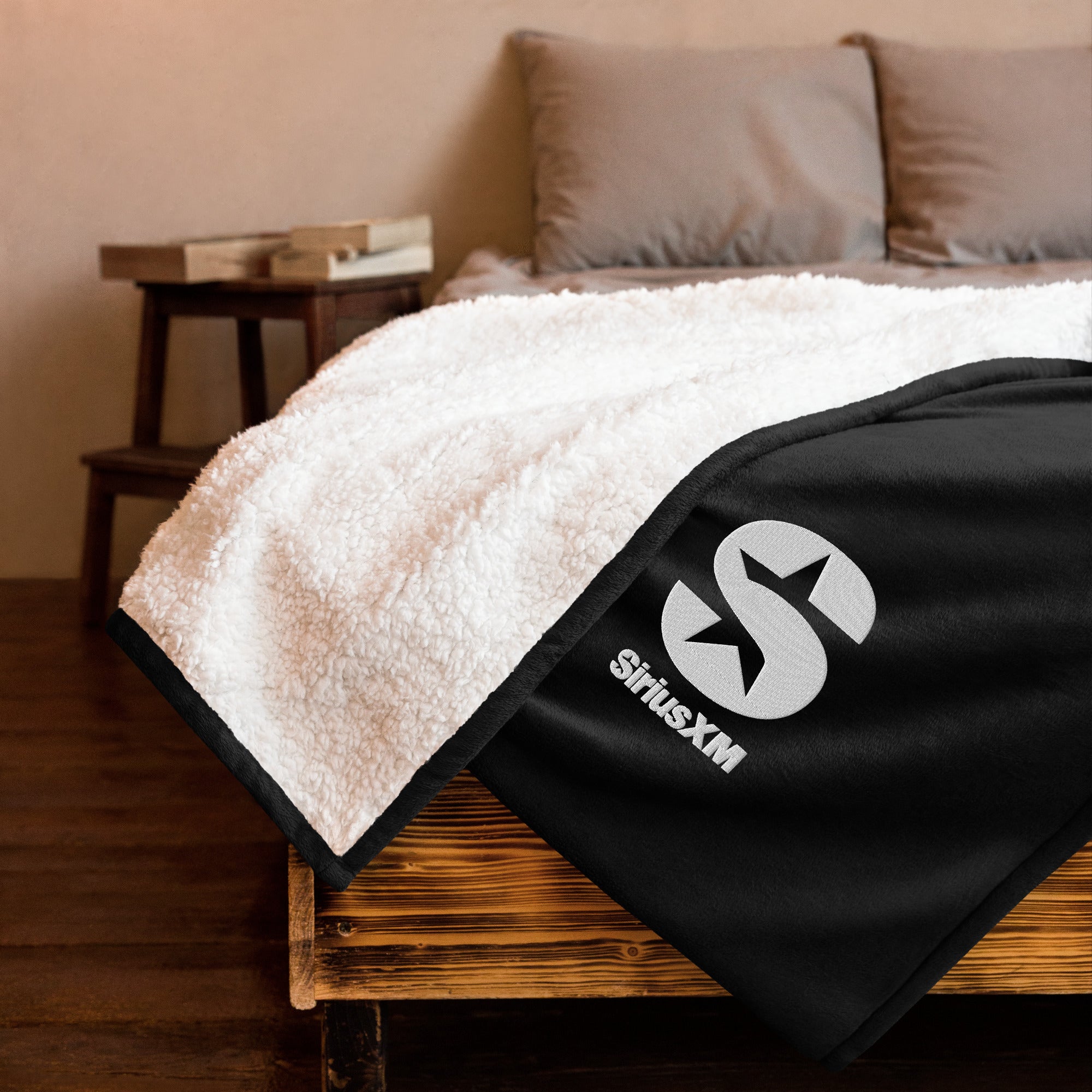 Cozy blanket featuring a black and white 'SiriusXM' logo, laid over a wooden bed with soft pillows in the background.