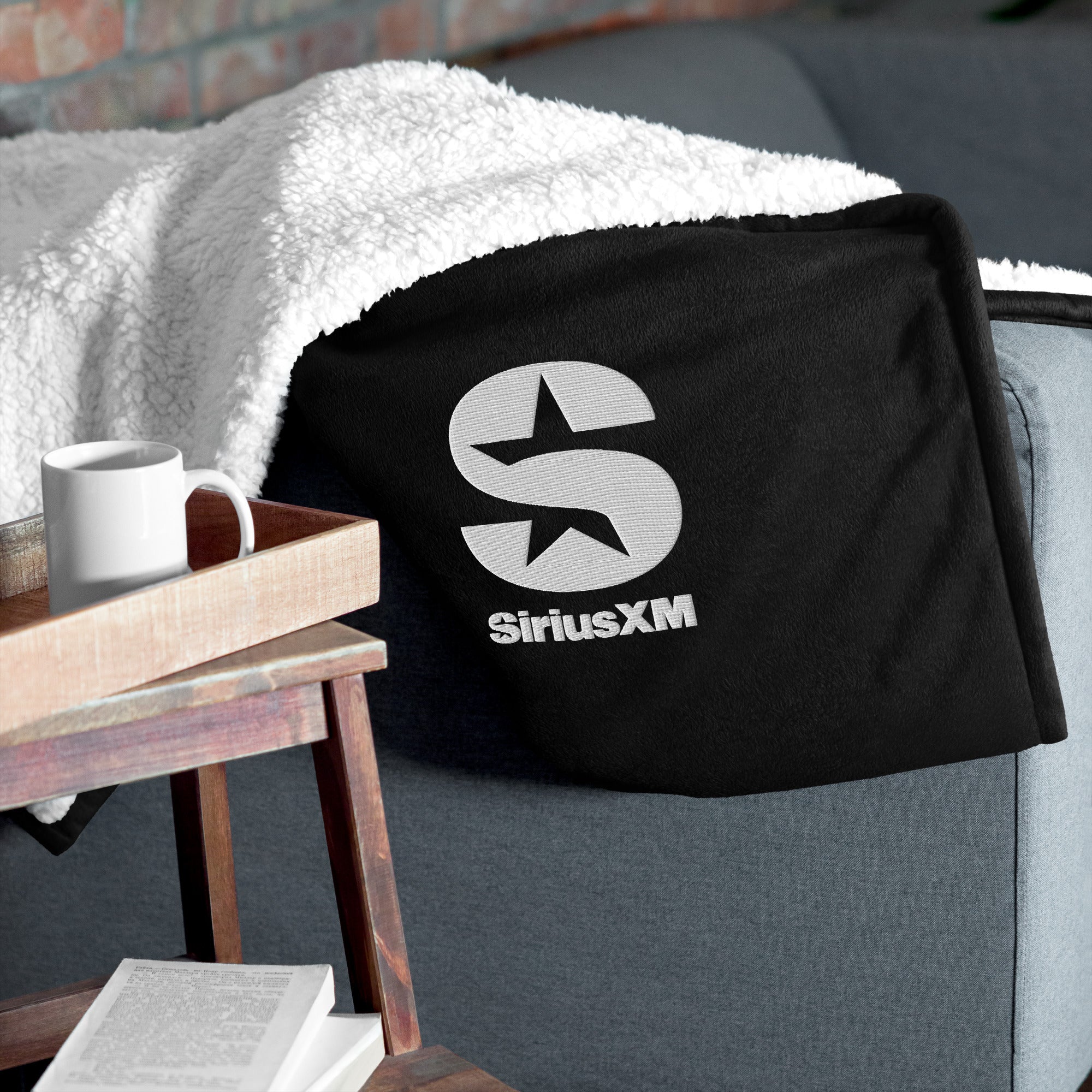 Black blanket with 'SiriusXM' logo draped over a blue couch, next to a wooden tray and mug. Cozy setting.