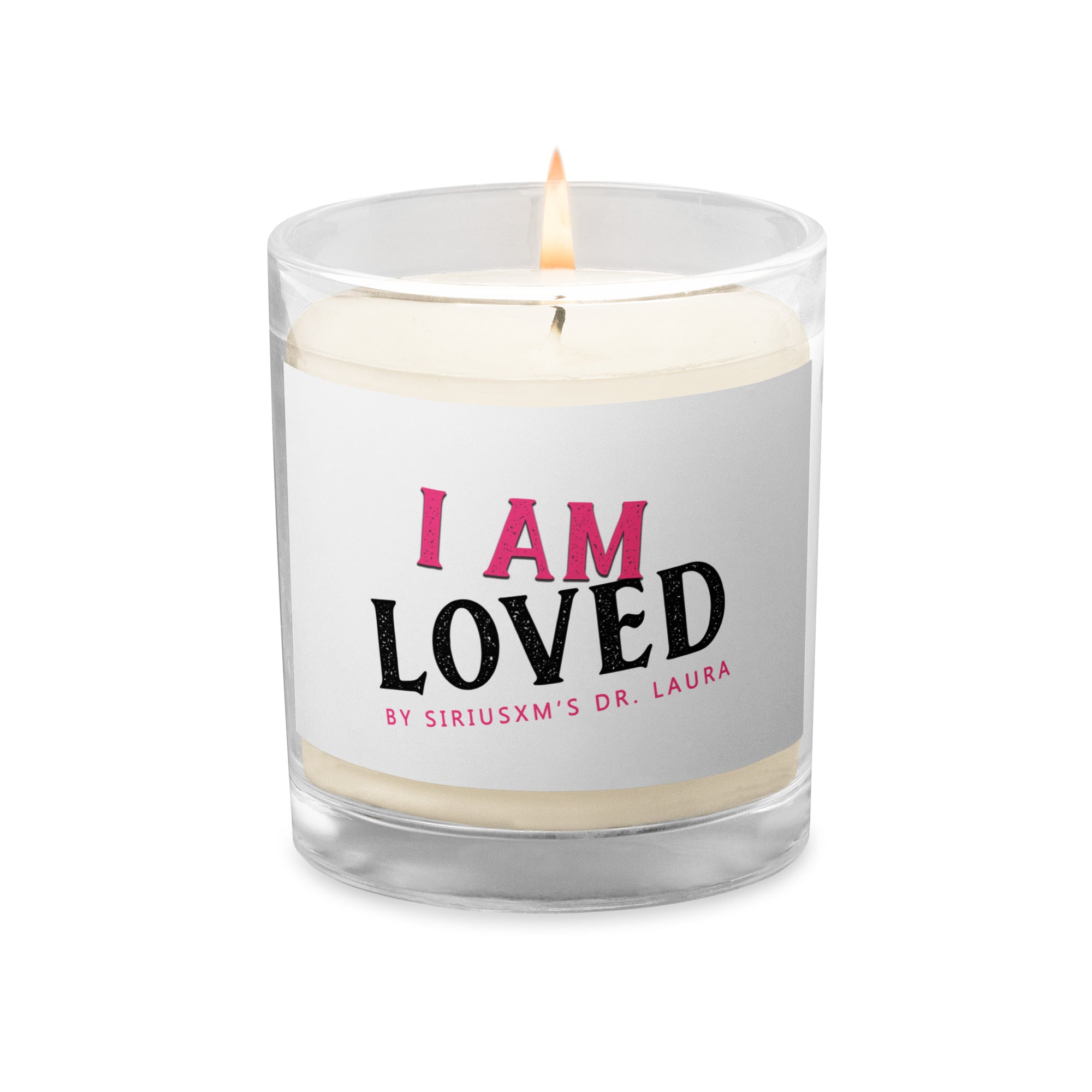 A clear glass jar candle with white soy wax inside. The label on the front reads 'I AM LOVED' in bold text, with 'I AM' in pink and 'LOVED' in black. Below, it says 'BY SIRIUSXM'S DR. LAURA' in smaller pink text. The candle wick is lit, producing a small flame